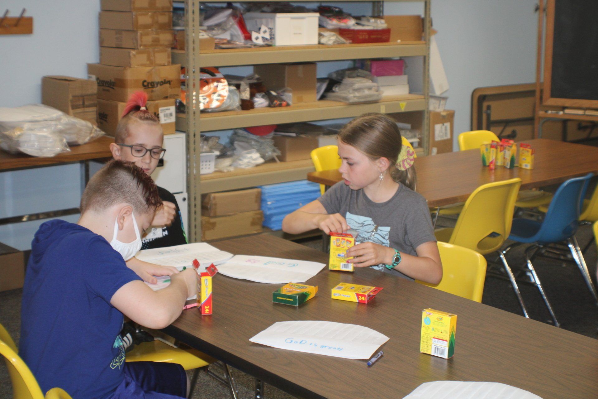 Children doing a table craft activity in a classroom, with shelves and yellow chairs in the background