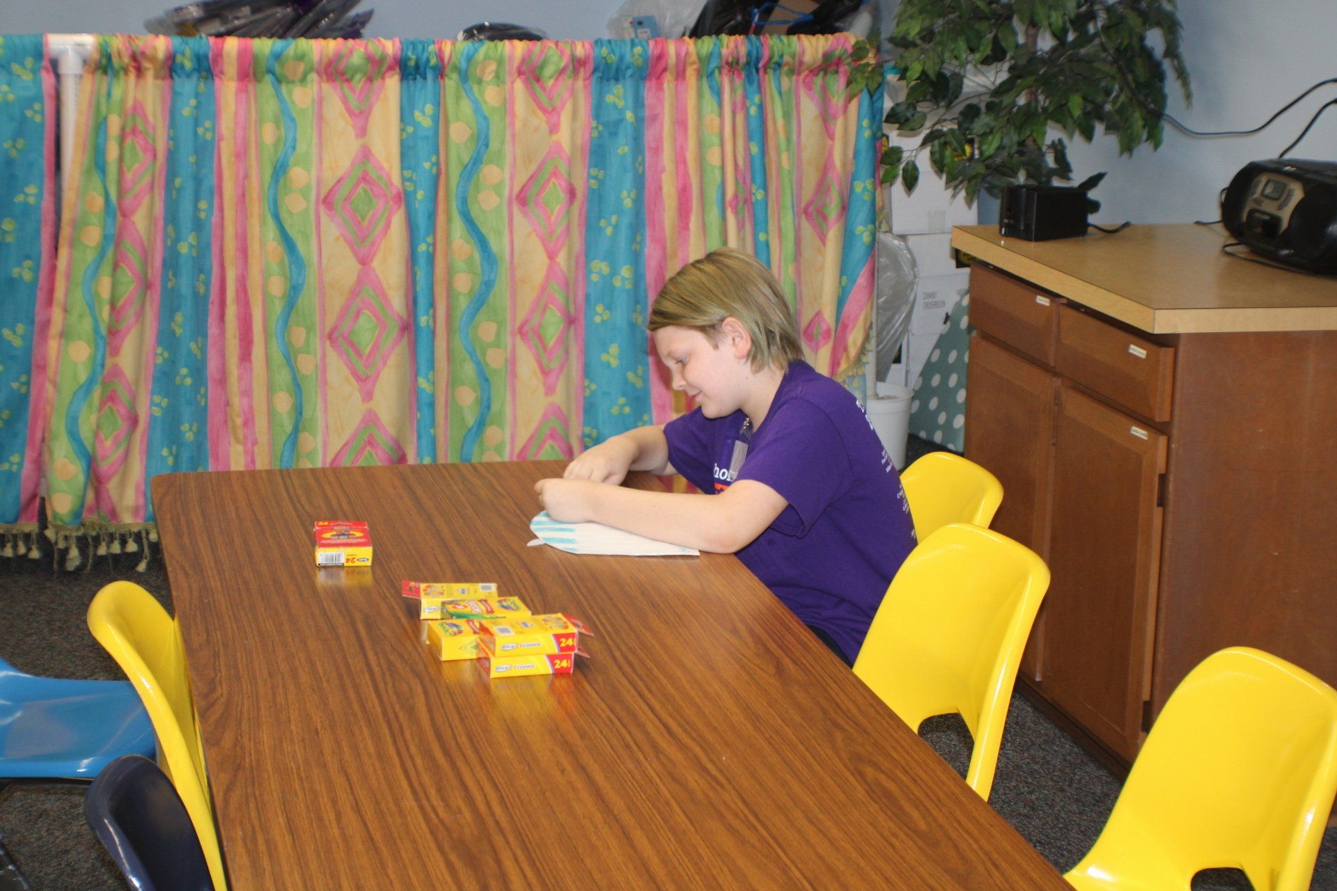 Person writing at a wooden table with colorful blocks in a room with bright yellow chairs and patterned curtains