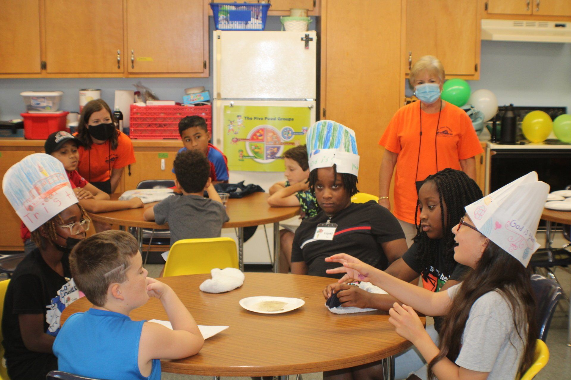 Children in a classroom sharing snacks at a round table, some wearing paper hats and costumes