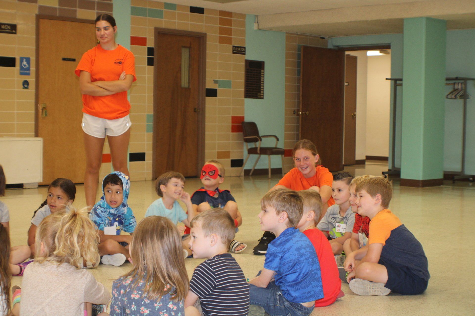 Children sitting on the floor in a lobby, listening to two adults in orange shirts.
