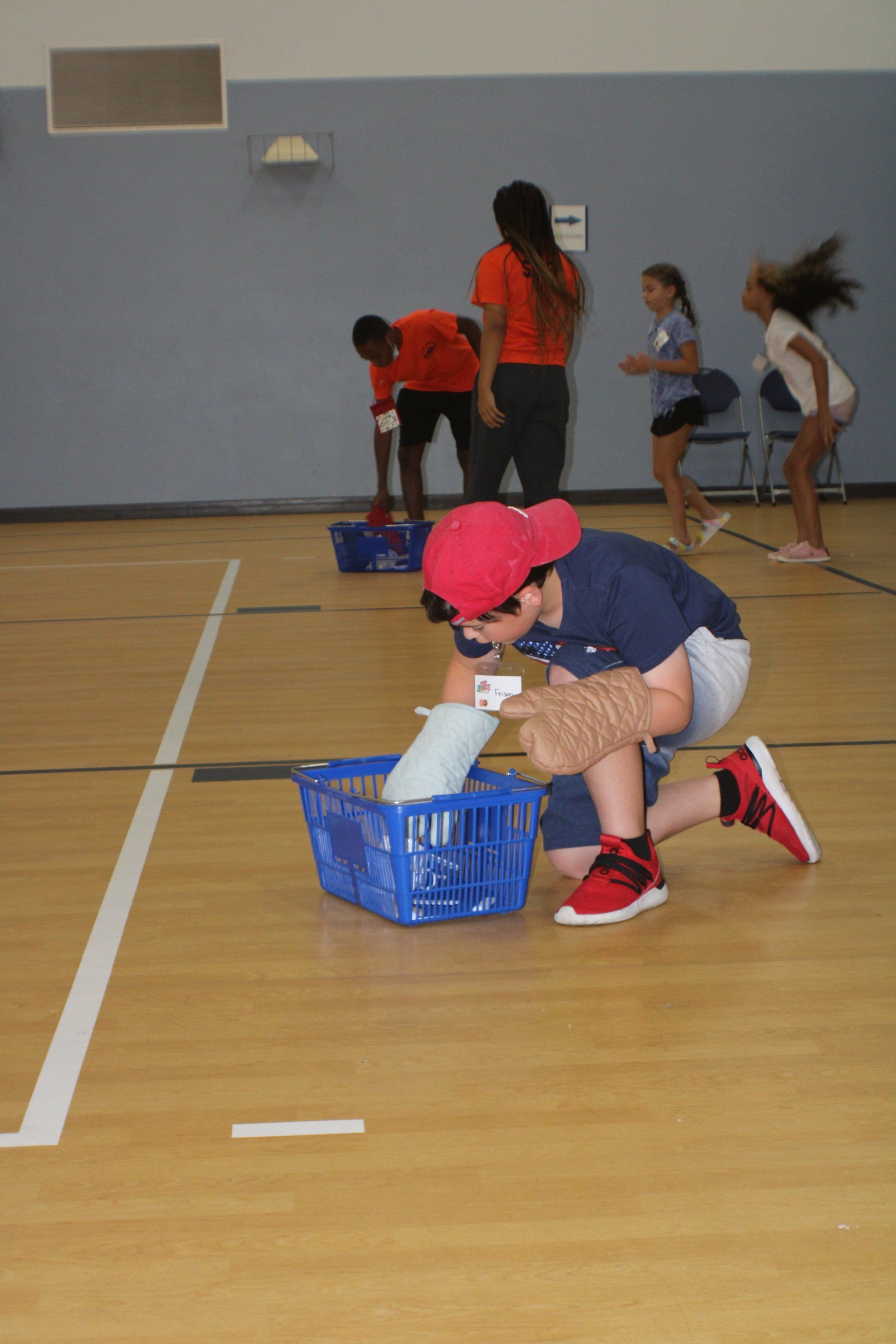 Children crouch in a gym, sorting items into blue baskets during an activity.