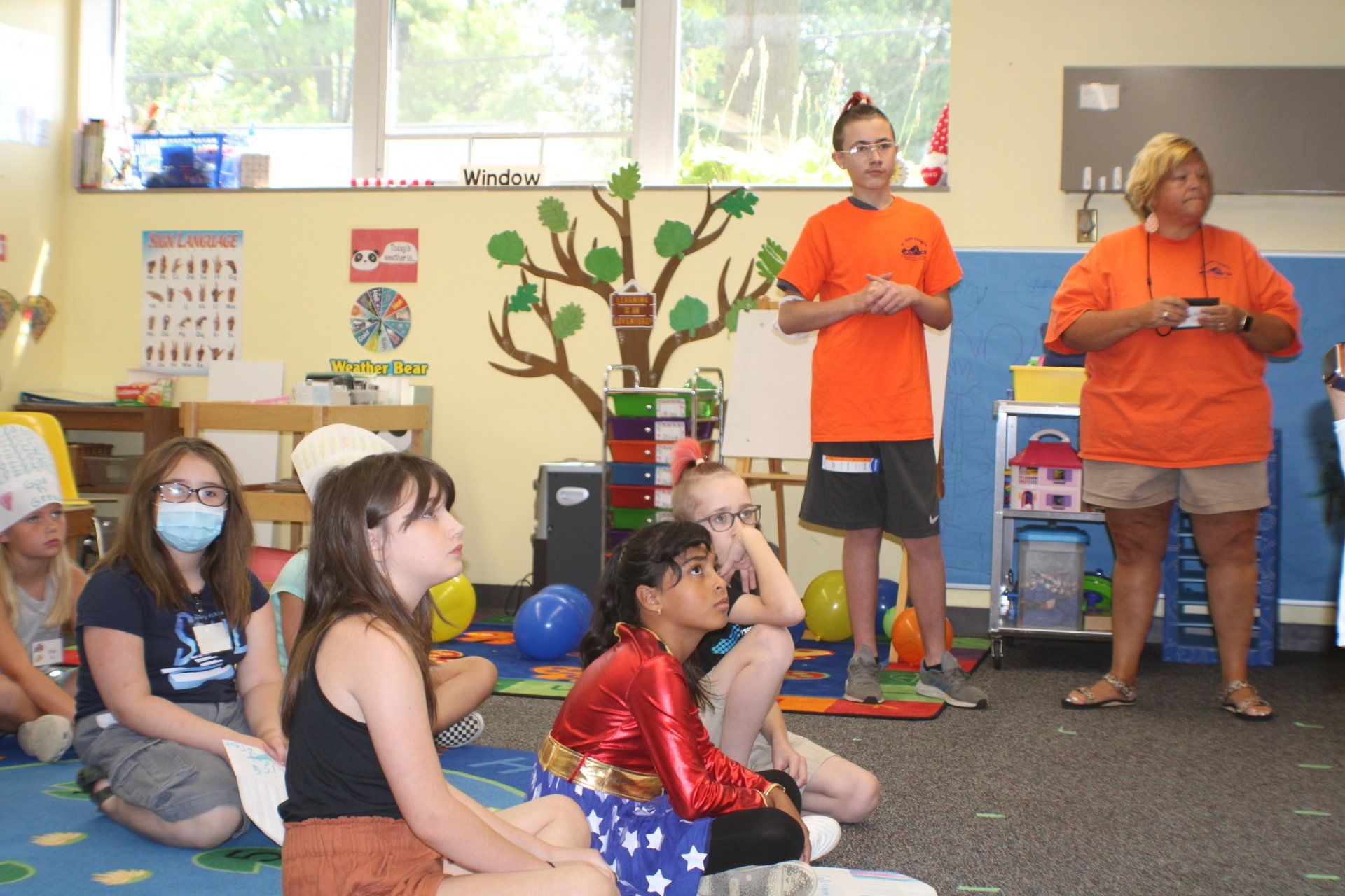 Children seated on a classroom carpet watching two adults in orange shirts speak near a tree display