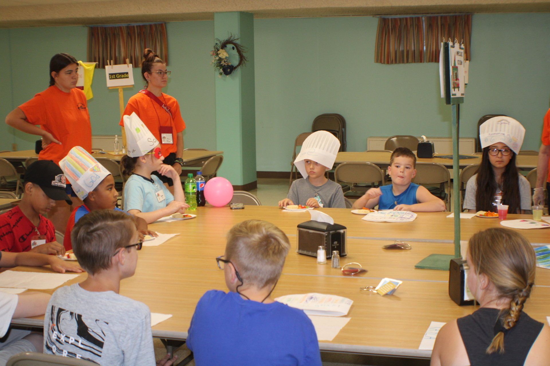 Children wearing paper hats sit around a table in a classroom, with two adults standing nearby.