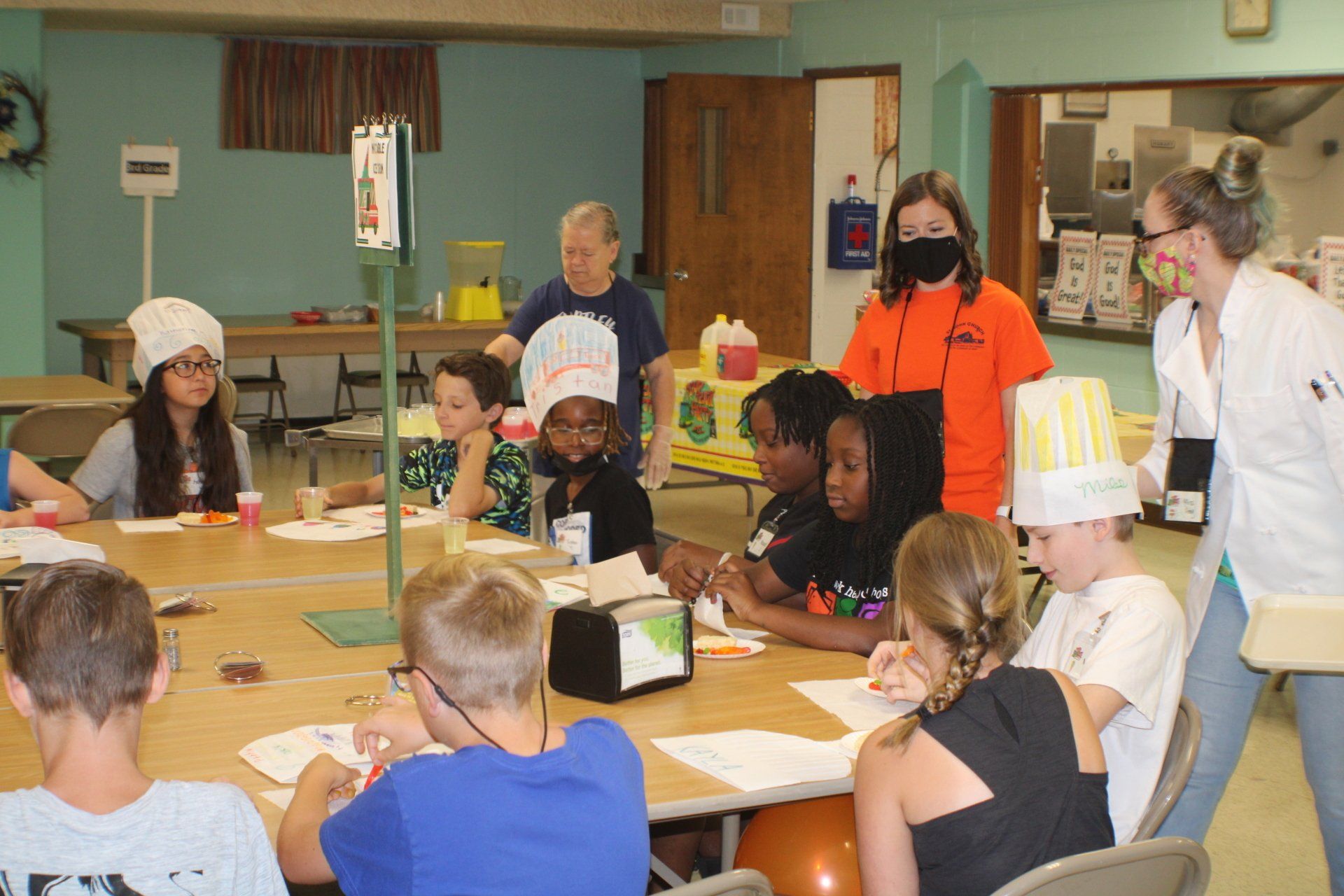 People gathered around a table in a classroom, wearing aprons and chef hats, preparing food together.