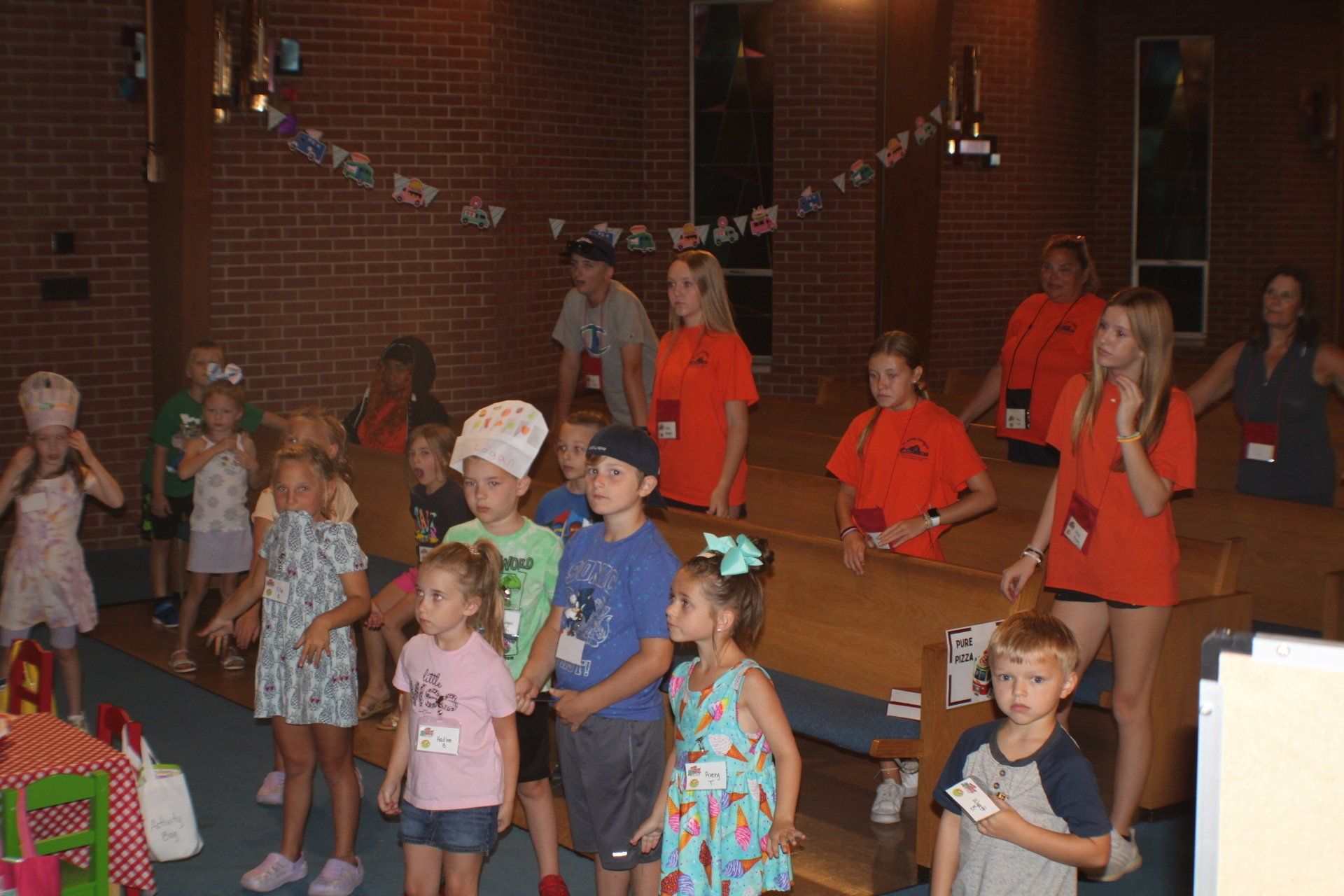 Children in colorful outfits performing on a stage in a brick-walled hall, with bunting overhead