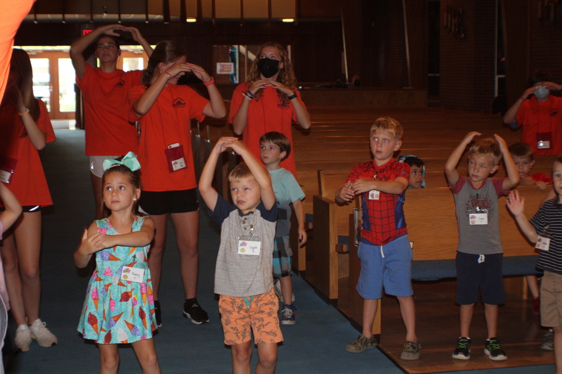 Children and adults in a hall doing a synchronized arm exercise with hands raised overhead
