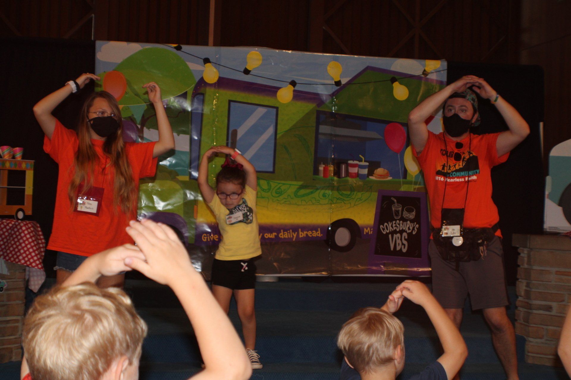 Three performers in colorful costumes pose onstage in front of a painted backdrop, while audience hands rise in front.