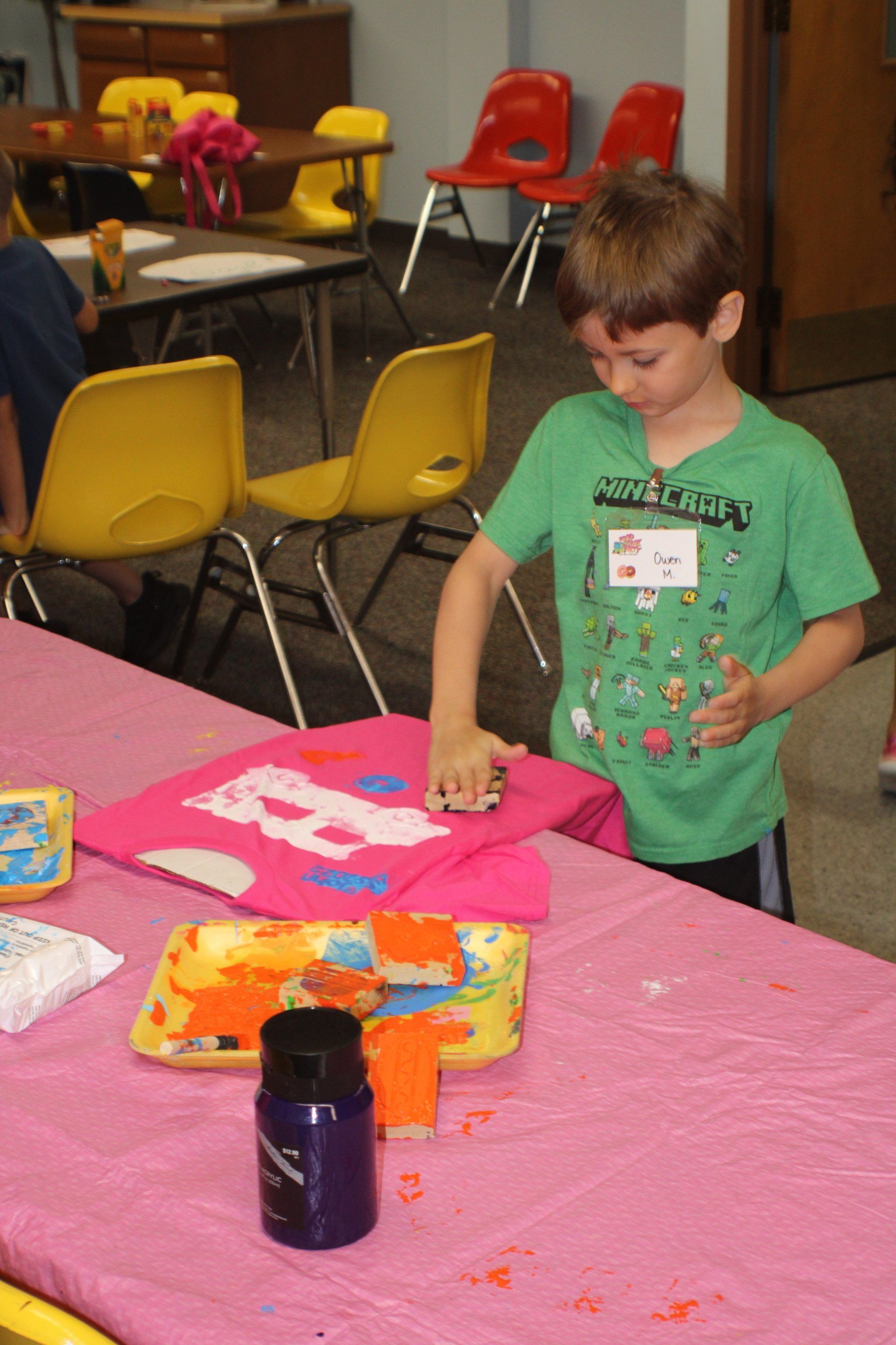 Child leaning over a pink-covered table, playing with colorful craft materials in a classroom.