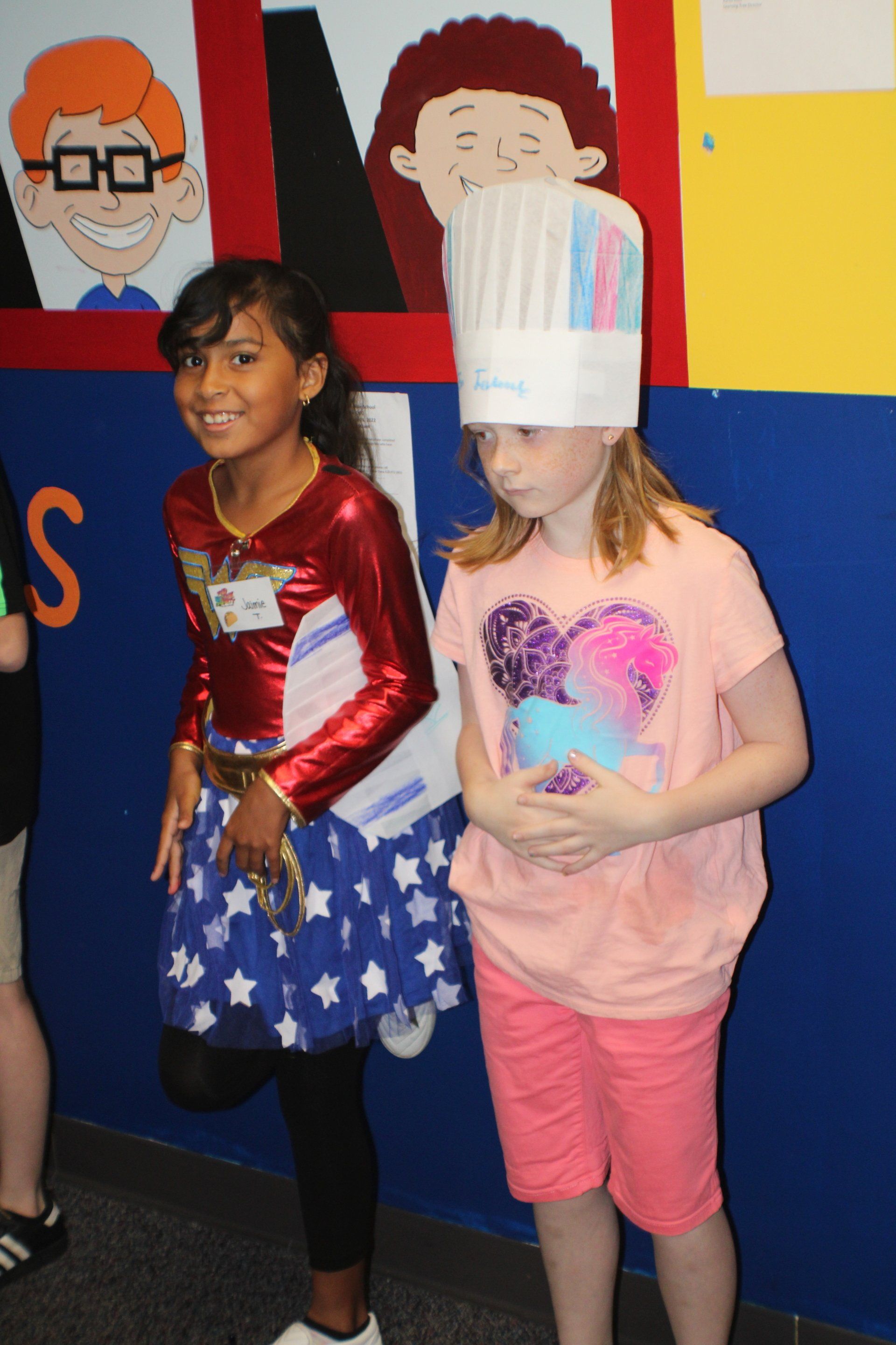 Two children standing indoors, one in a red top and star skirt, the other in a pink outfit with a paper hat.