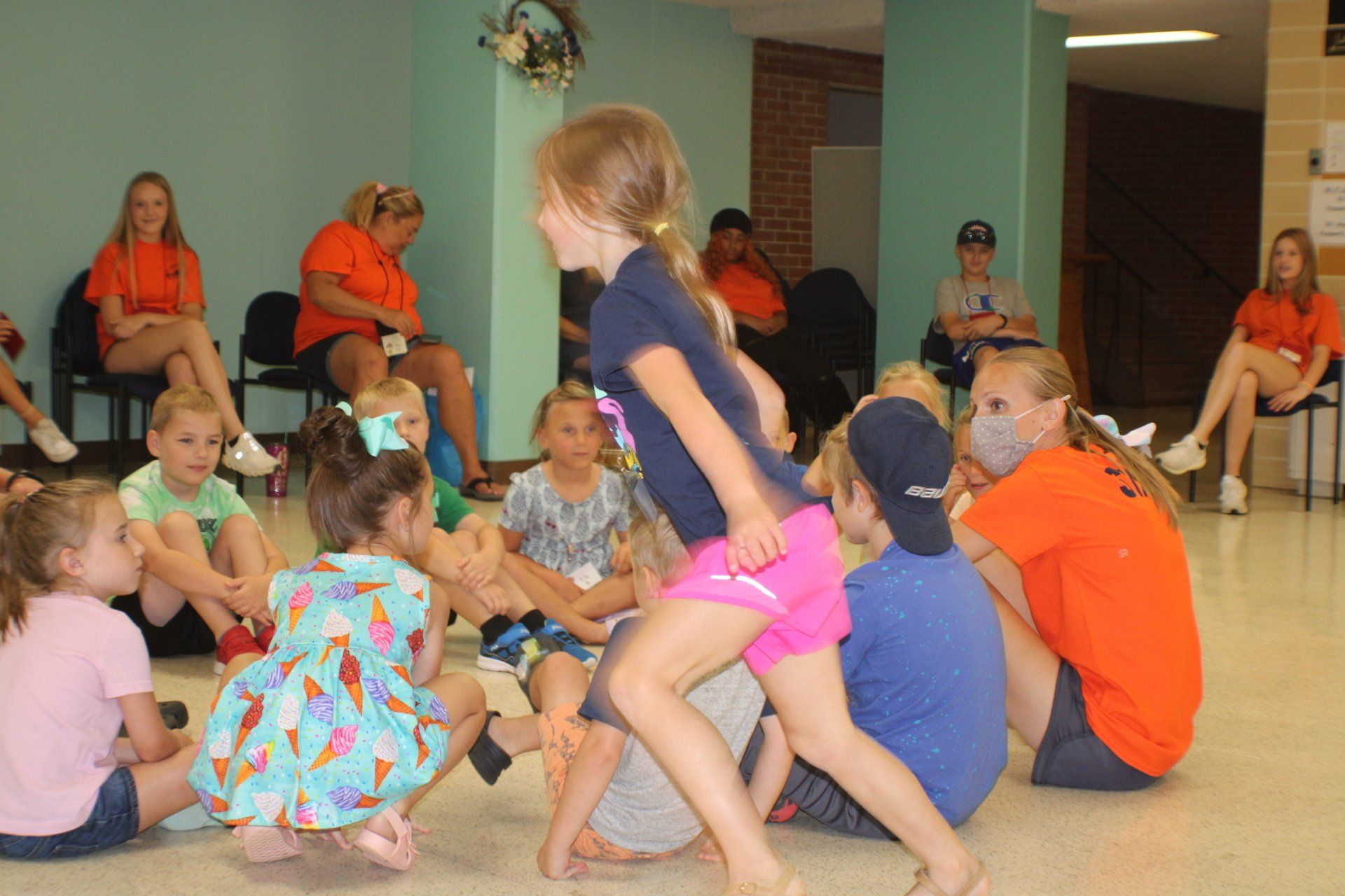 Children sit and play in a gym, with one child in pink shorts standing in the center.