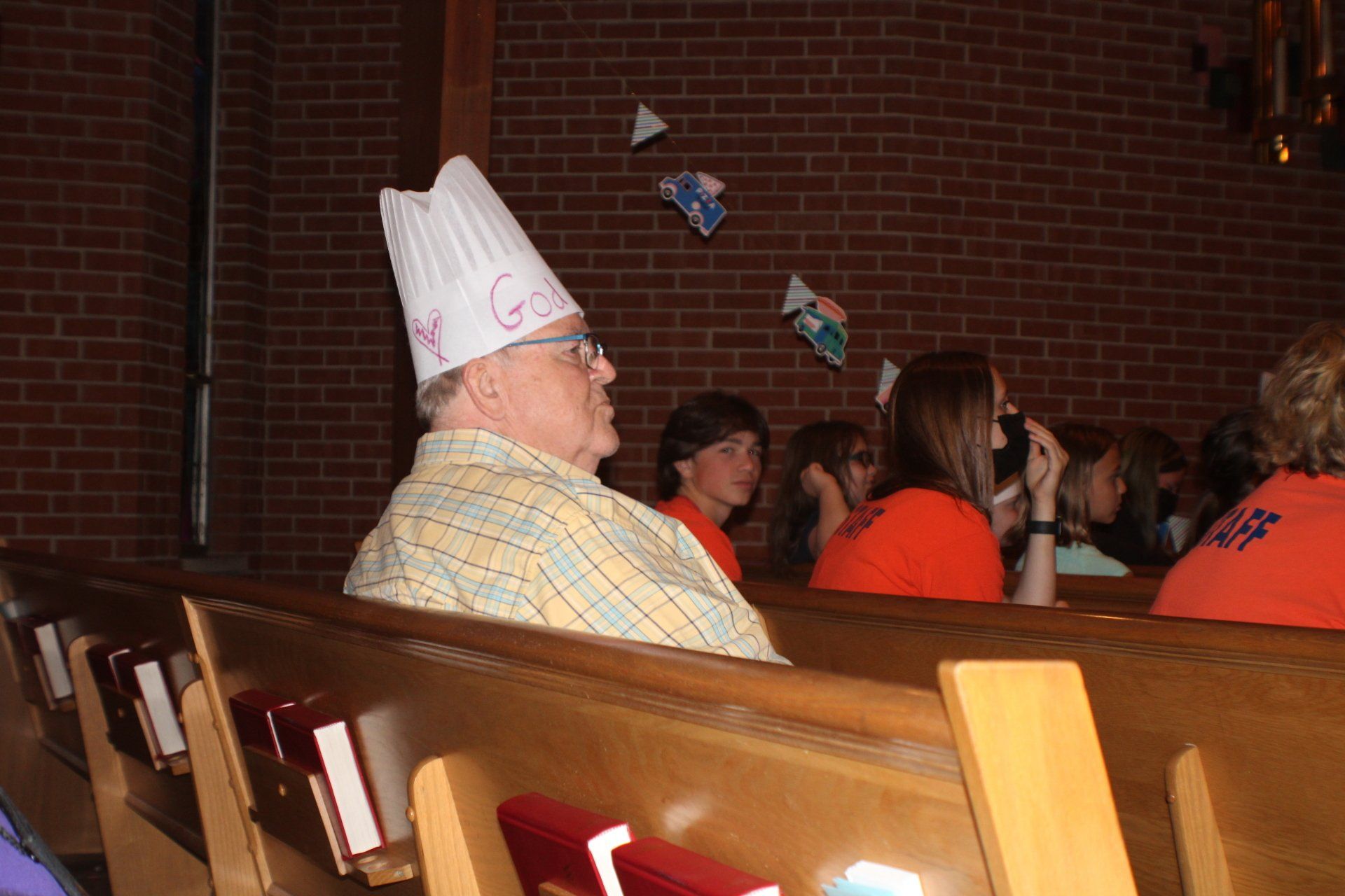 Person in a paper hat seated at a church event with others in orange shirts and paper decorations overhead