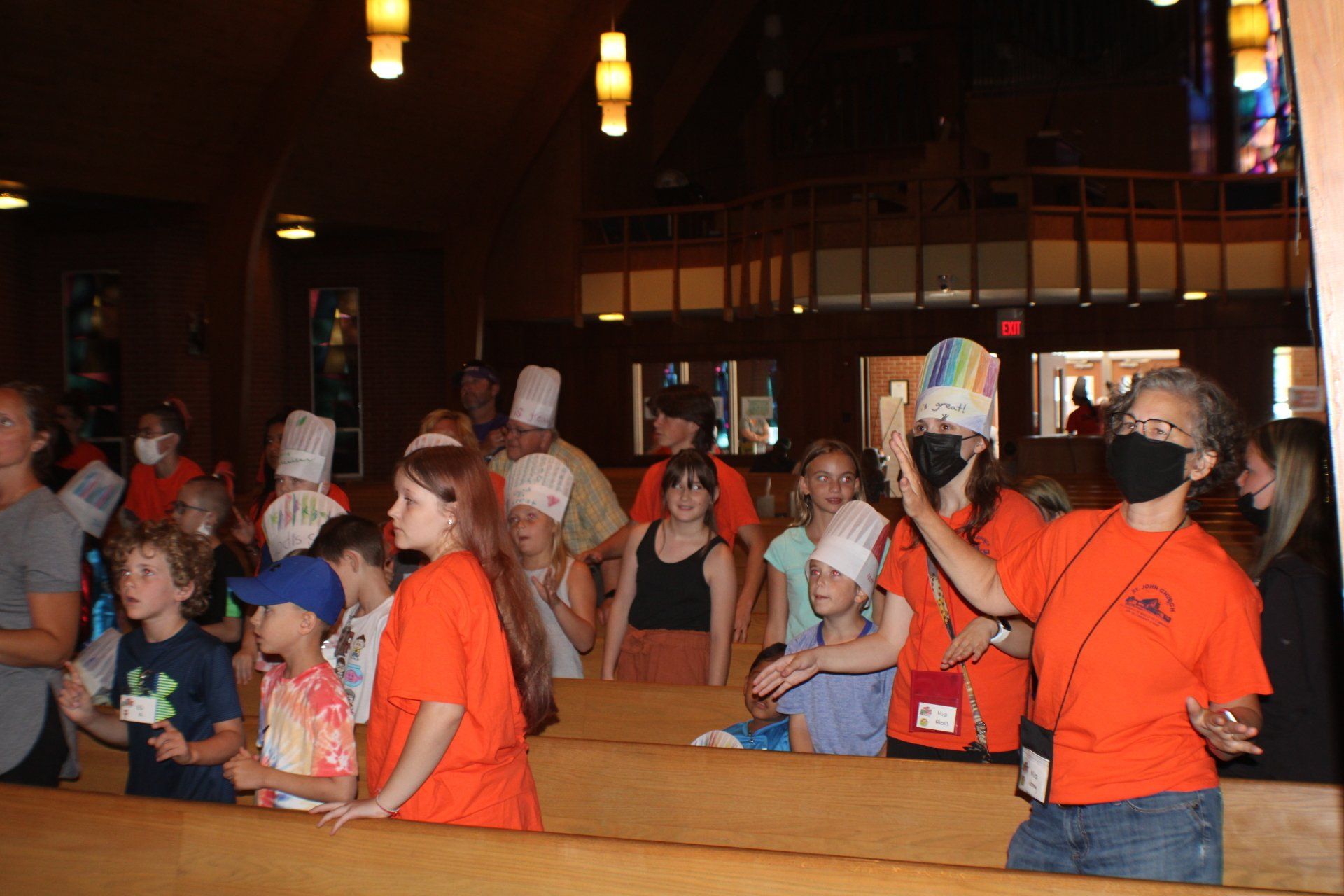 Children in paper hats and orange shirts seated in a dim restaurant, raising hands and chatting