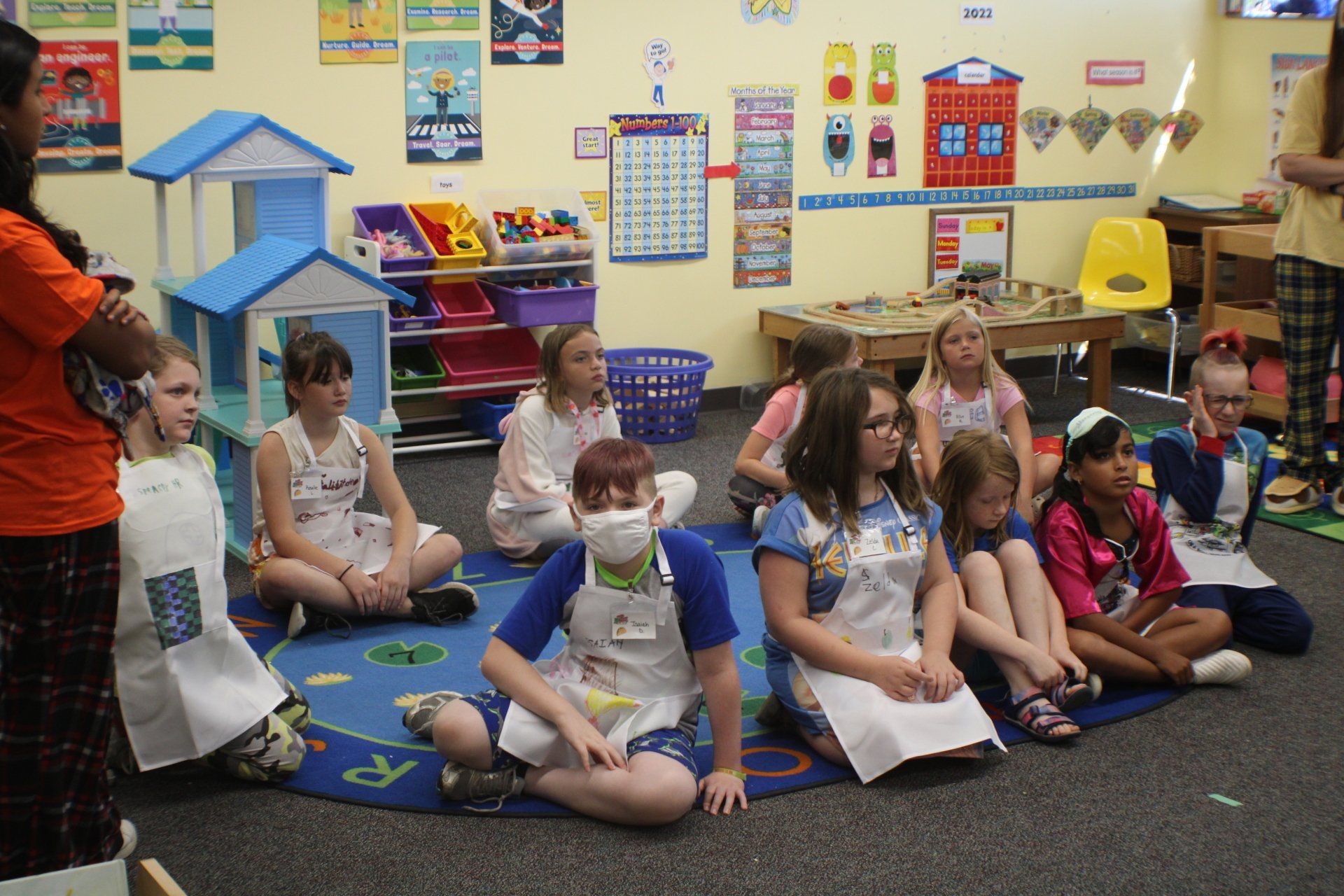 Children sitting on a classroom carpet during a group activity, with colorful posters and learning materials behind them