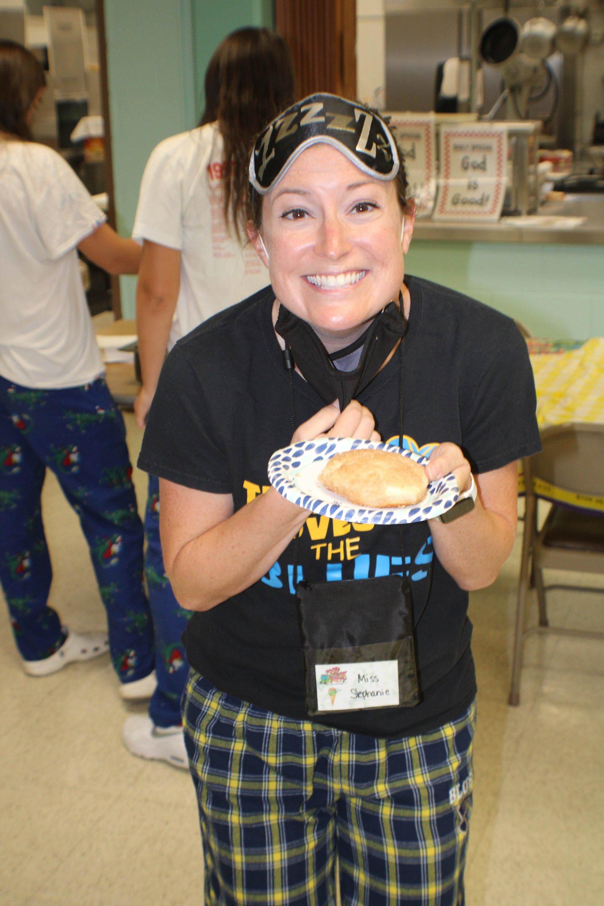 Person smiling and holding a plate of food indoors, wearing a black shirt and plaid pants.
