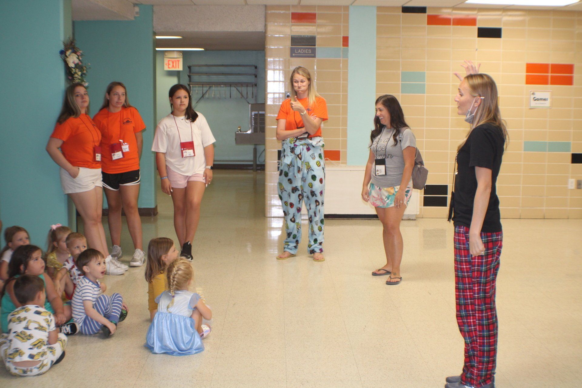 Teacher speaking to children seated on a school hallway floor while other adults stand nearby.