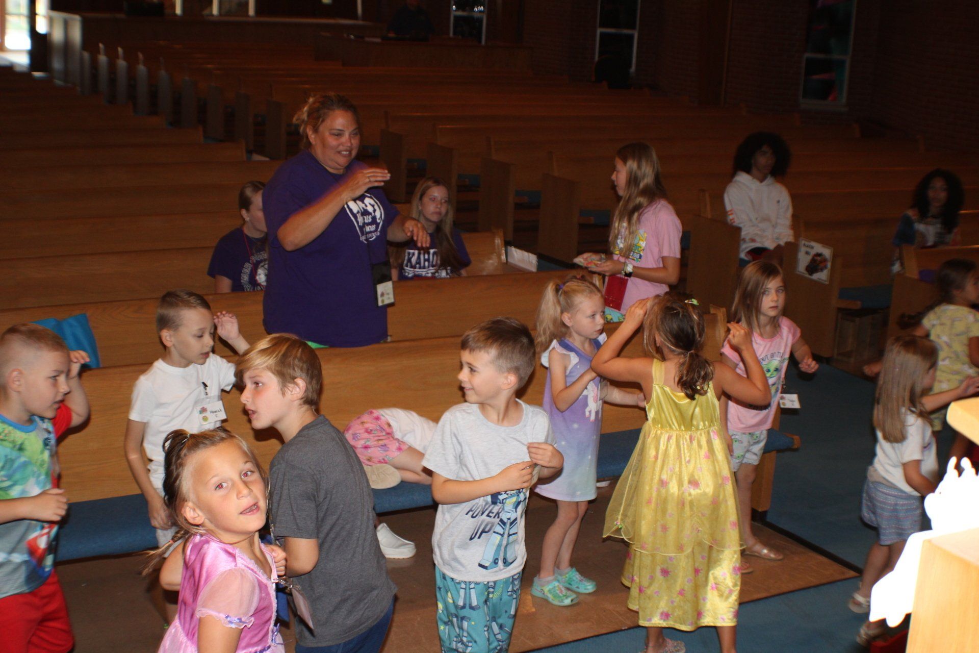 Children in colorful costumes and a woman stand in a wooden hall, clapping and dancing.