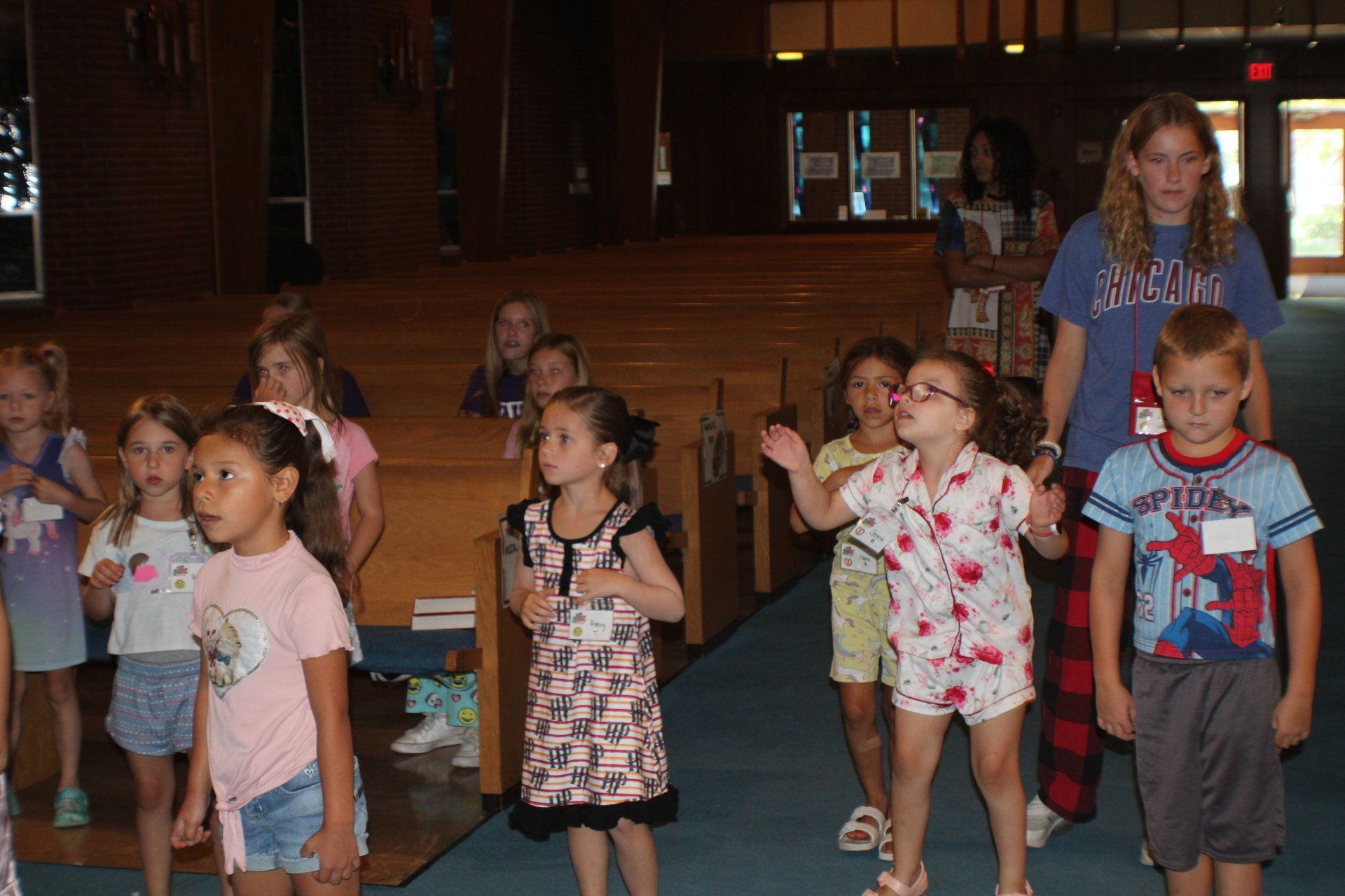 Children standing and waving in a dim auditorium, with a woman behind them