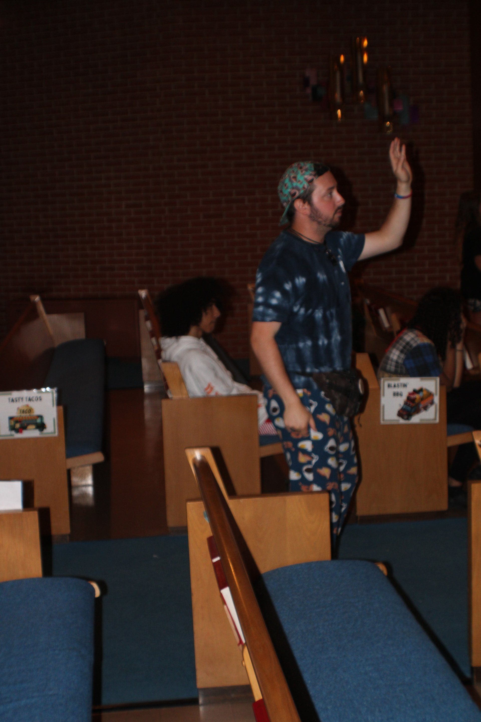 Person standing in a dim church pew area, raising a hand during a service.