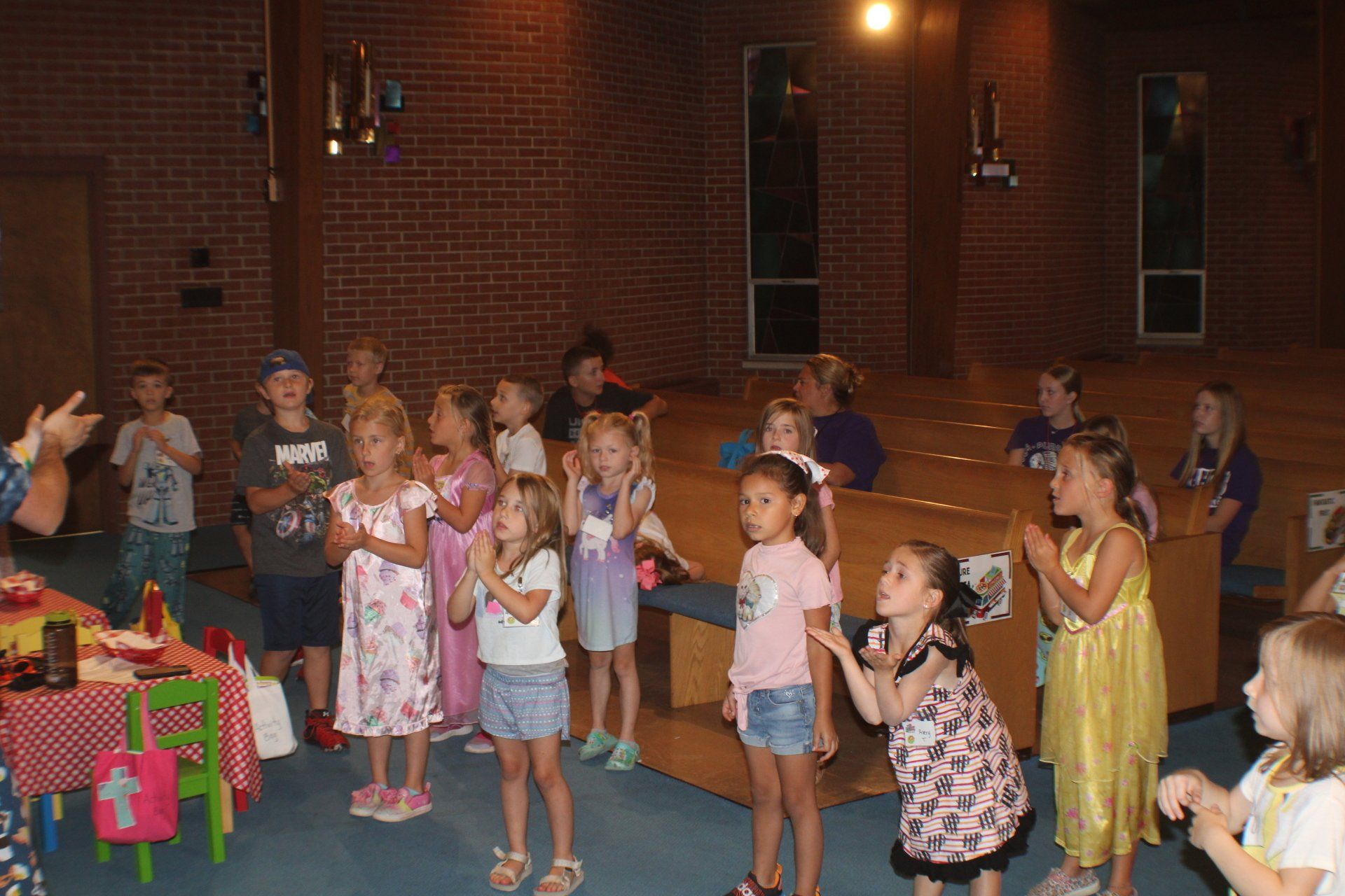 Children standing in a line, clapping and singing in a gym beside colorful gift boxes.