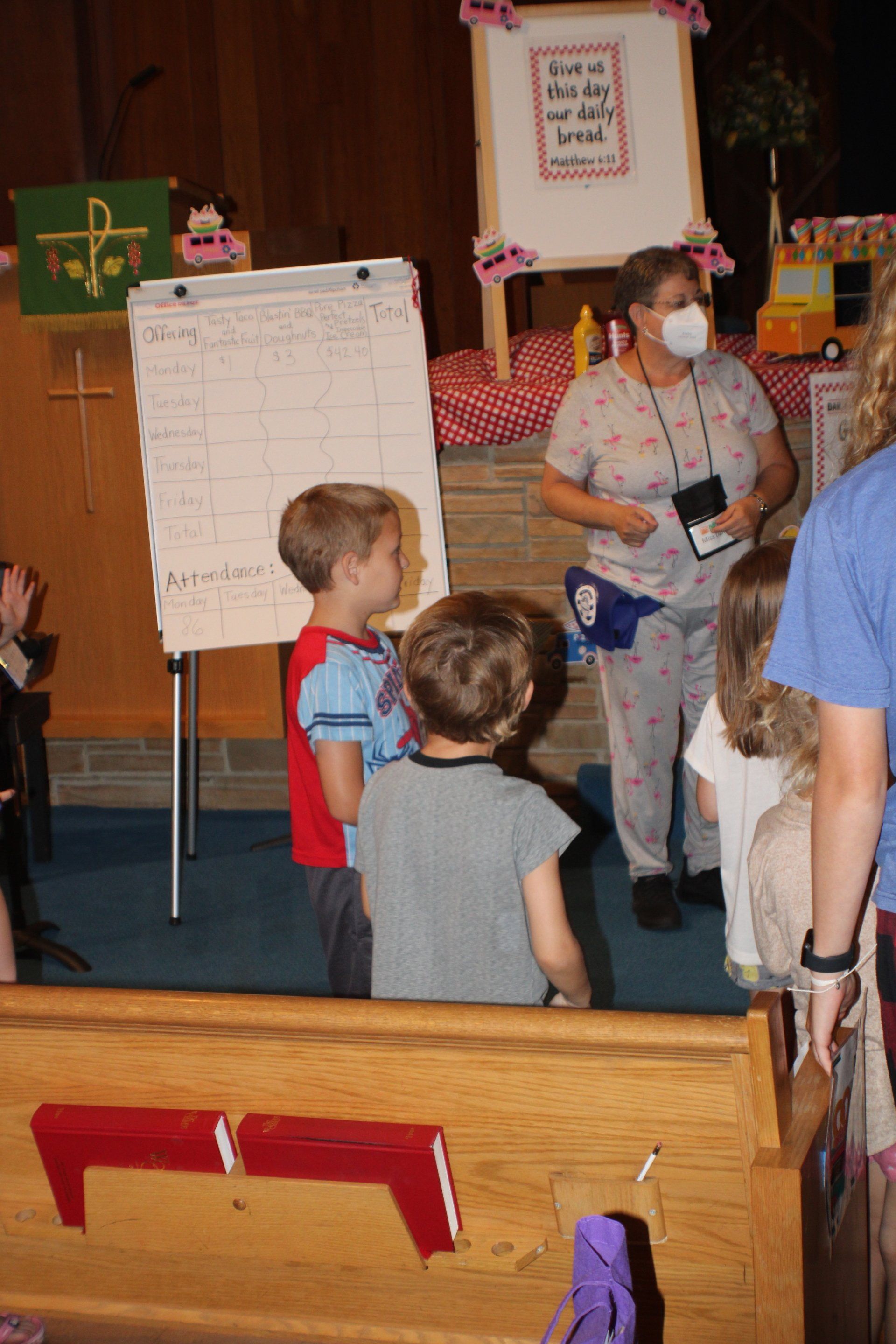 Children and an adult in a classroom, with a whiteboard and pink decorations.