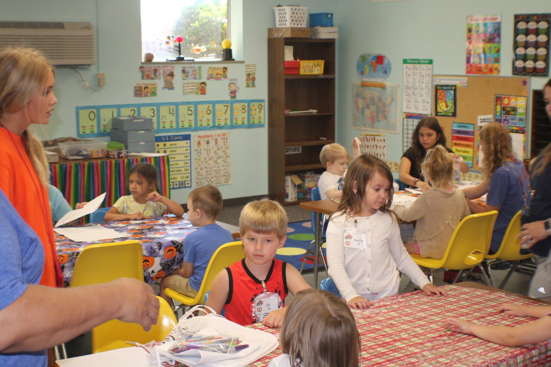 Children sitting at tables in a colorful classroom while a teacher stands at the front