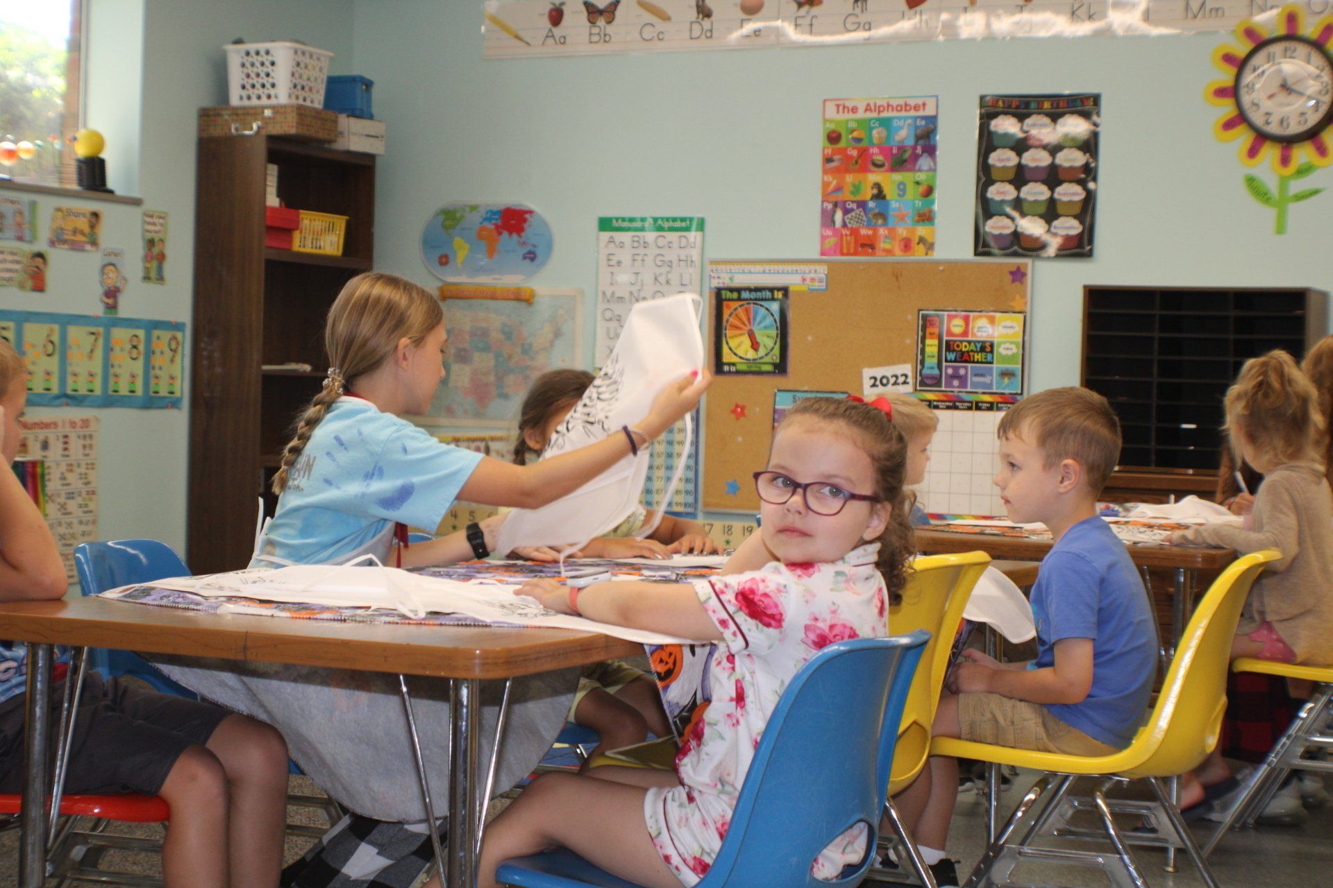Children working at tables in a colorful classroom, with a teacher assisting them.