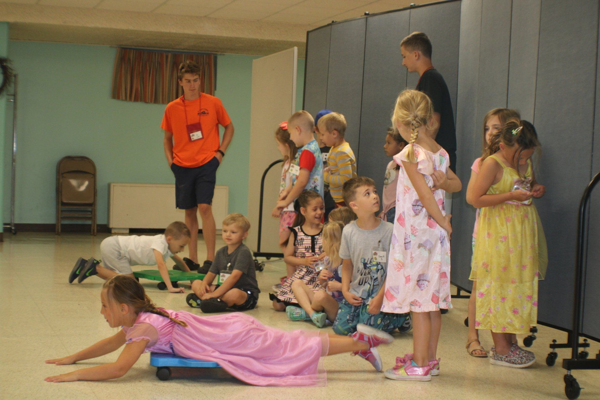 Children in colorful costumes on a gym floor, with adults watching in the background