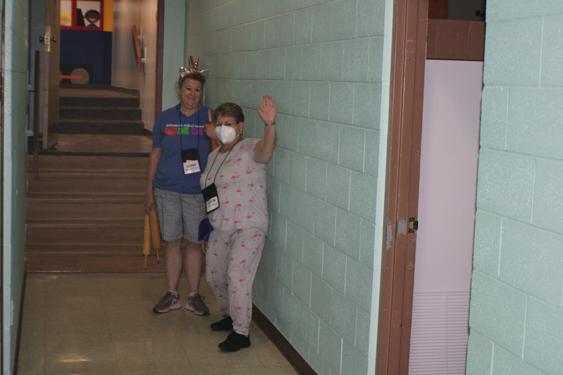 Two people stand in a hallway, one waving and wearing a mask.