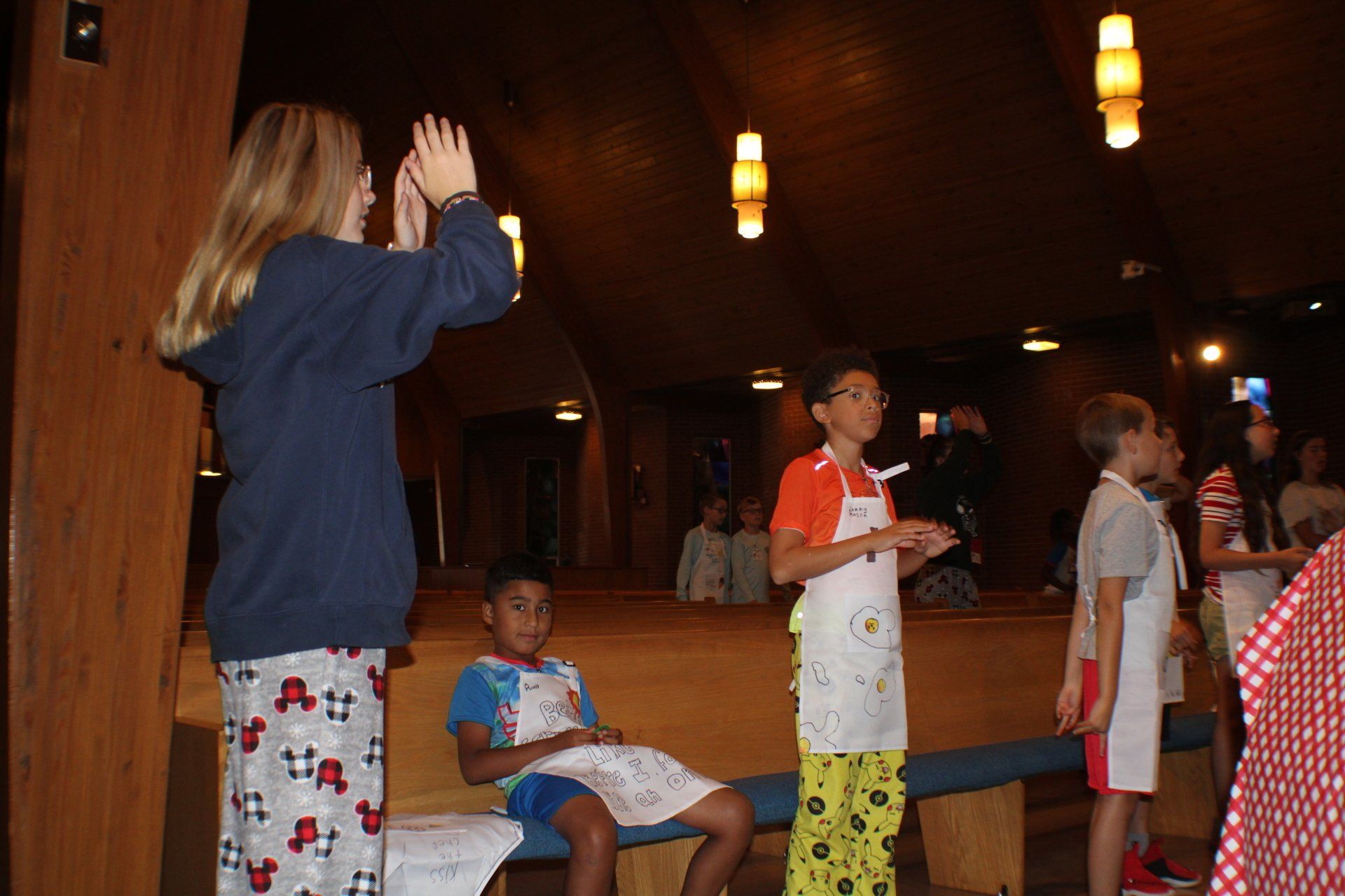 Children playing in a wood-paneled room with hanging lights; one child raises both hands nearby.
