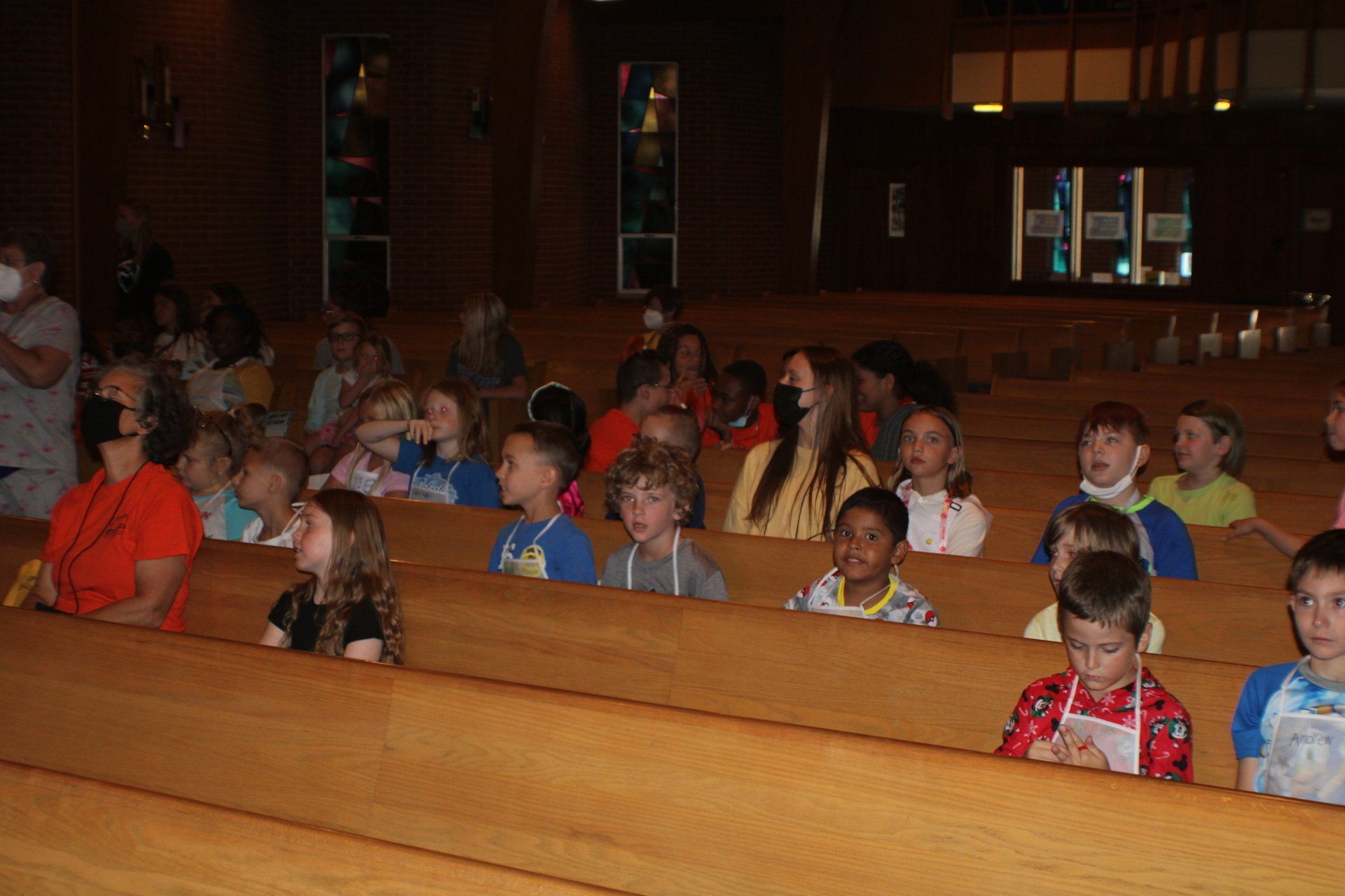 Children seated in wooden church pews during a gathering, some wearing face masks