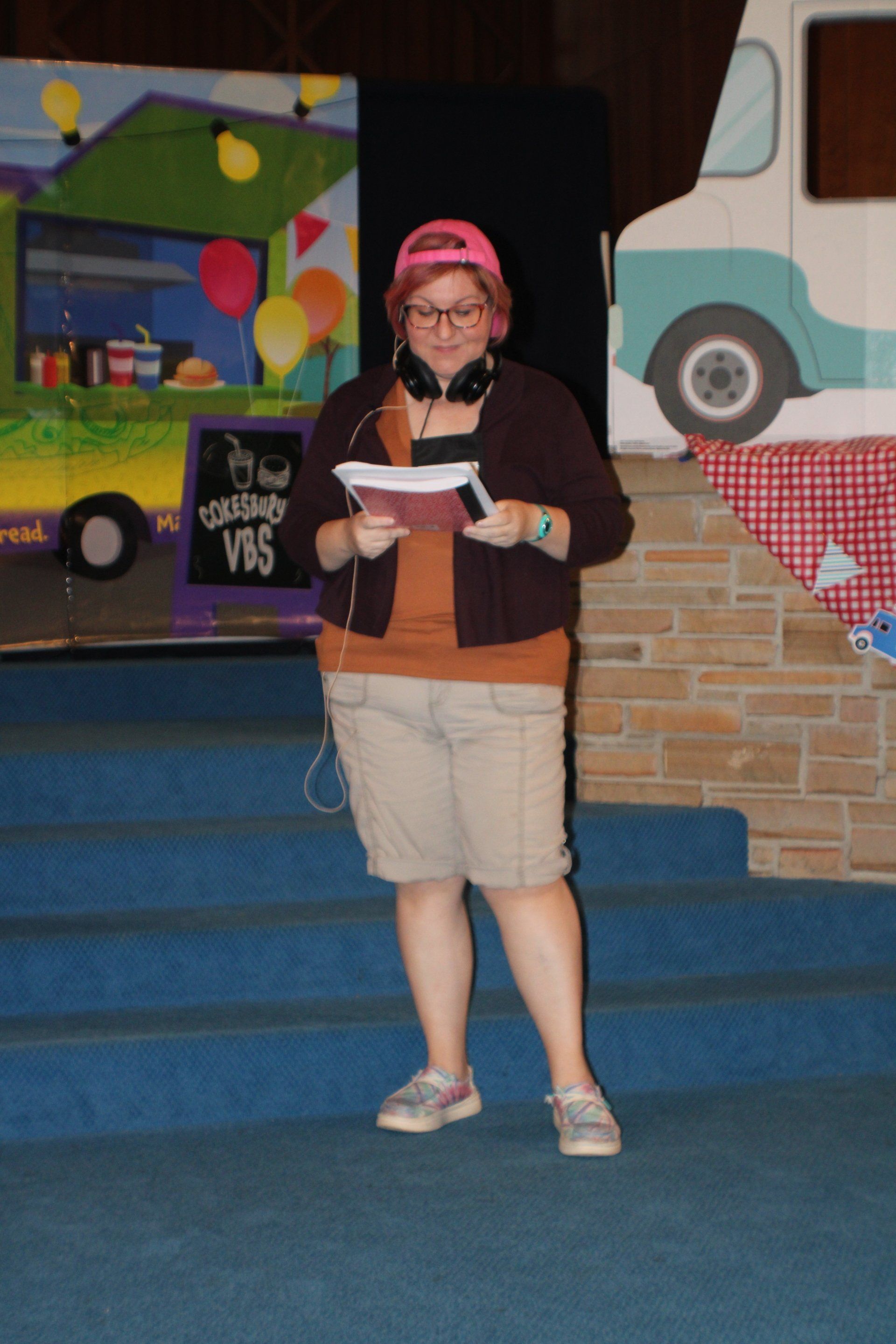 Person standing on a stage reading from papers, wearing a pink cap and tan shorts.