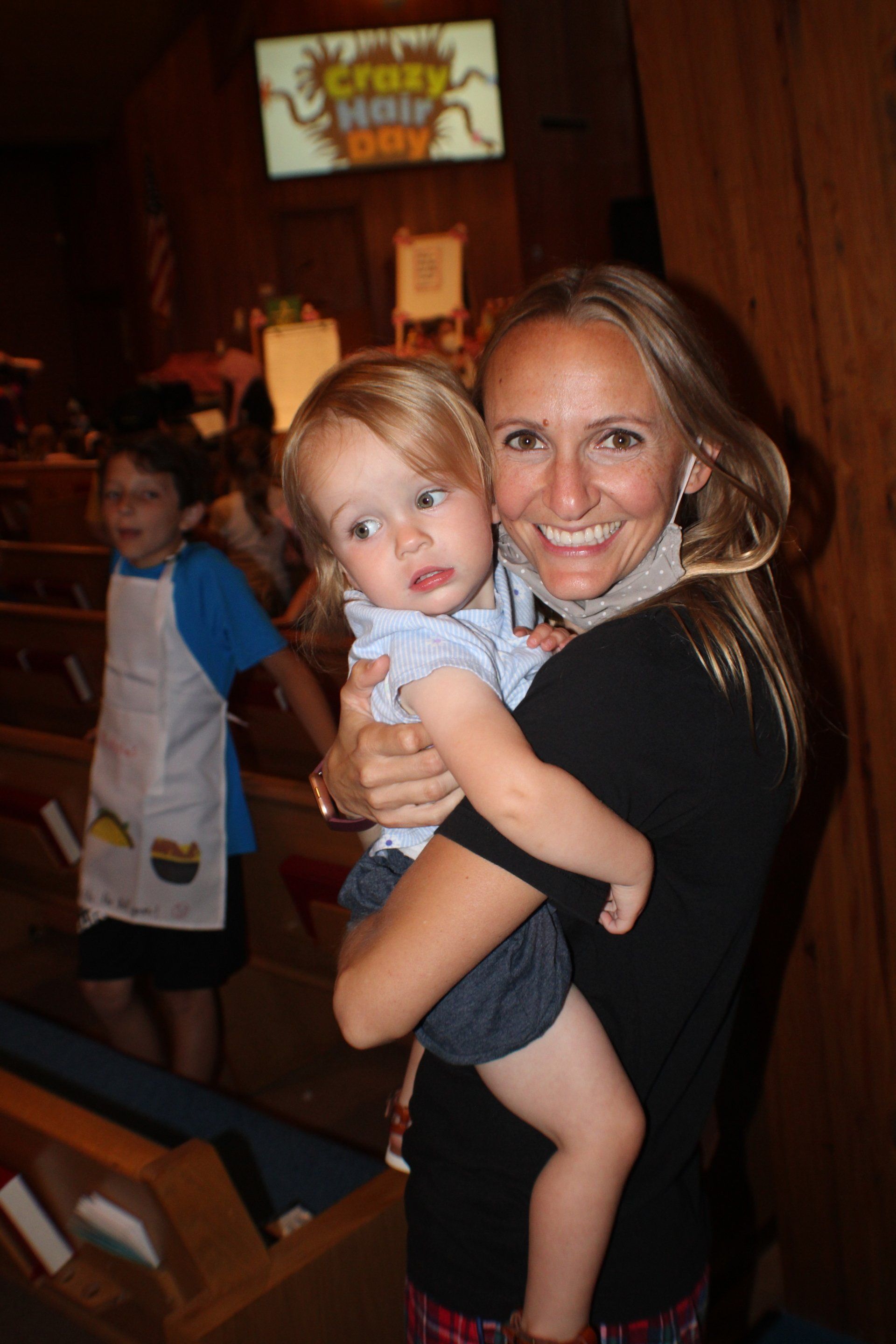 Smiling woman holding a toddler in a wooden church pew, with another child in the background.