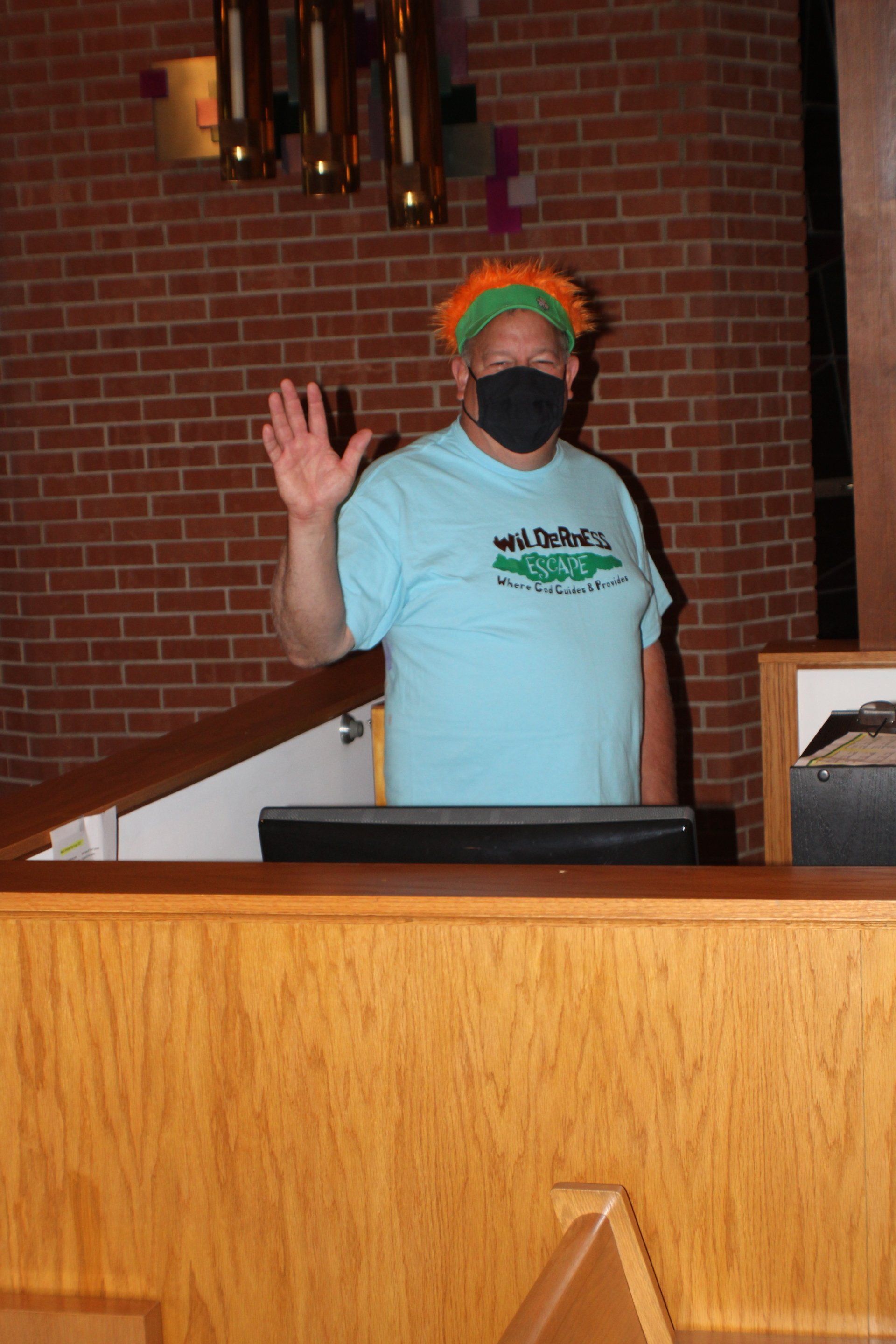Masked person in light blue shirt waving behind a wooden podium in a brick-walled room