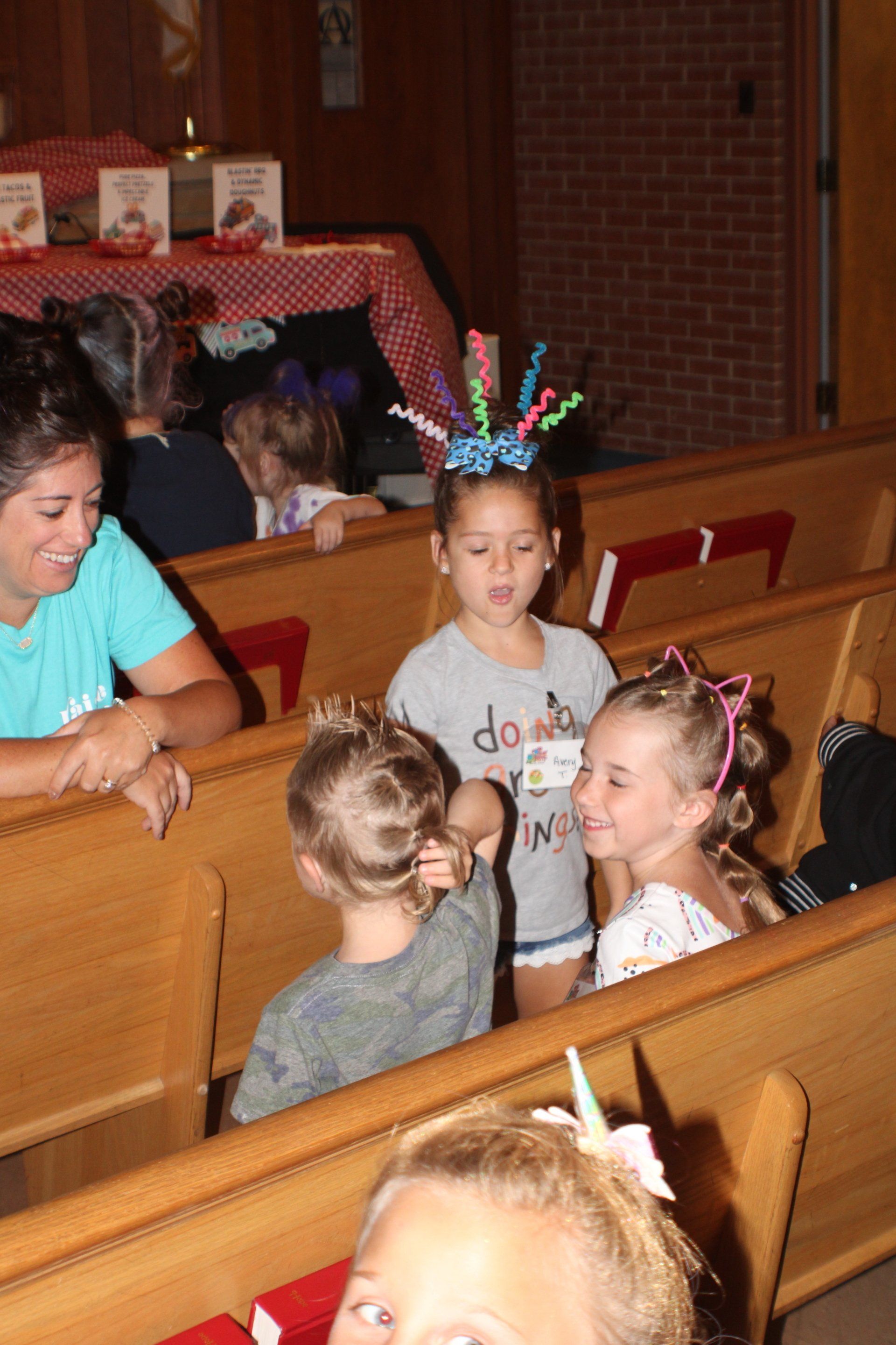 Children in church pews talking, with a woman seated nearby and colorful headbands visible.