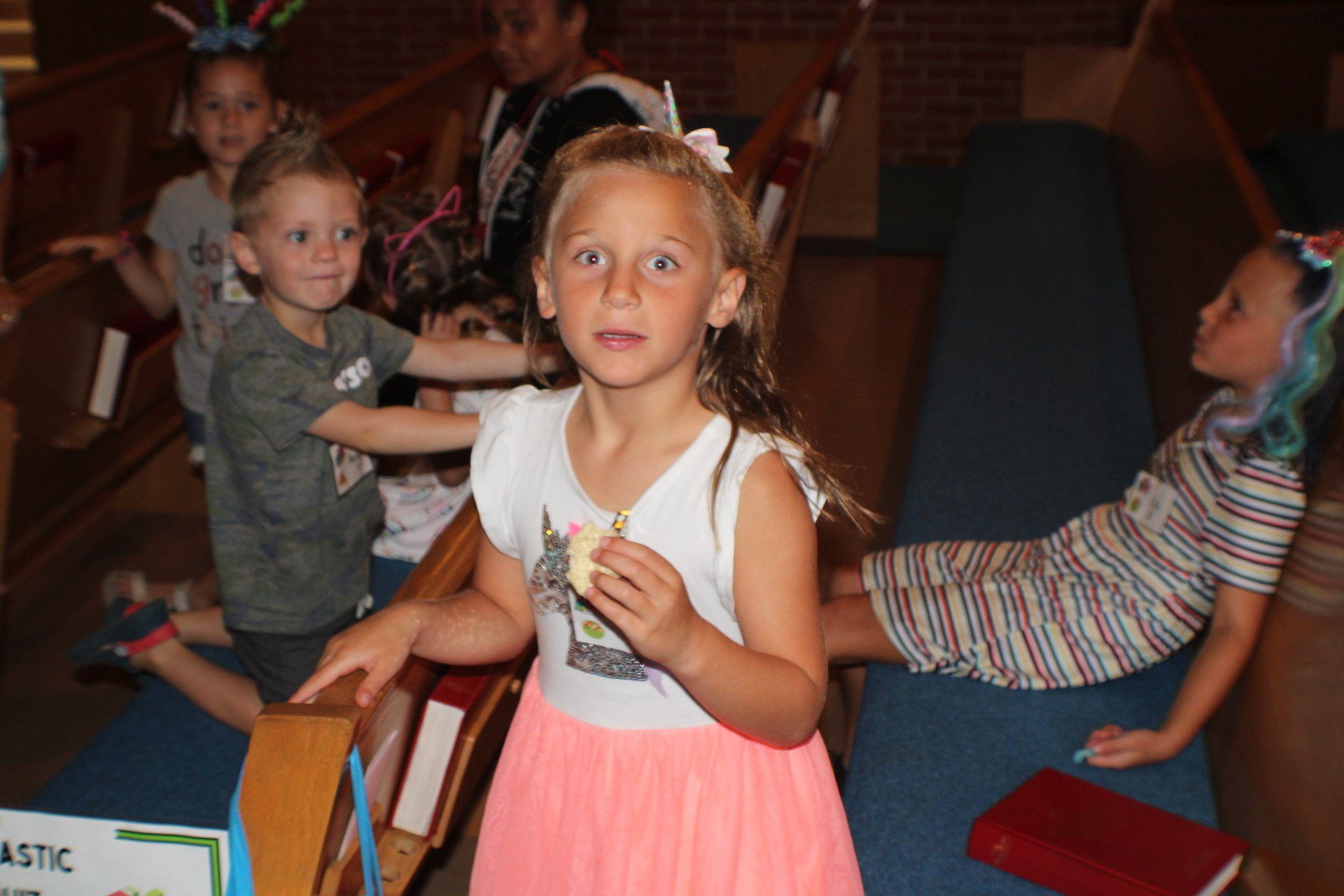 Children standing among chairs in a dim indoor room, one girl in a pink dress in the foreground