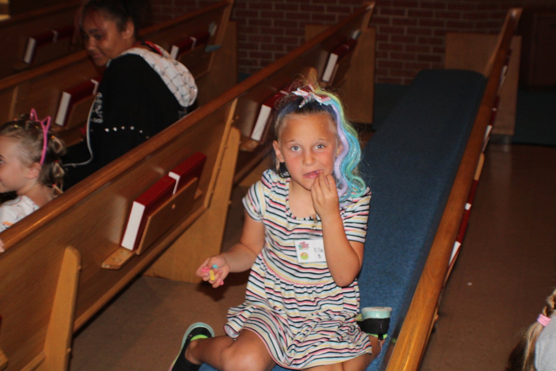 Child sitting in a church pew, wearing a striped dress and blue hair ribbons.