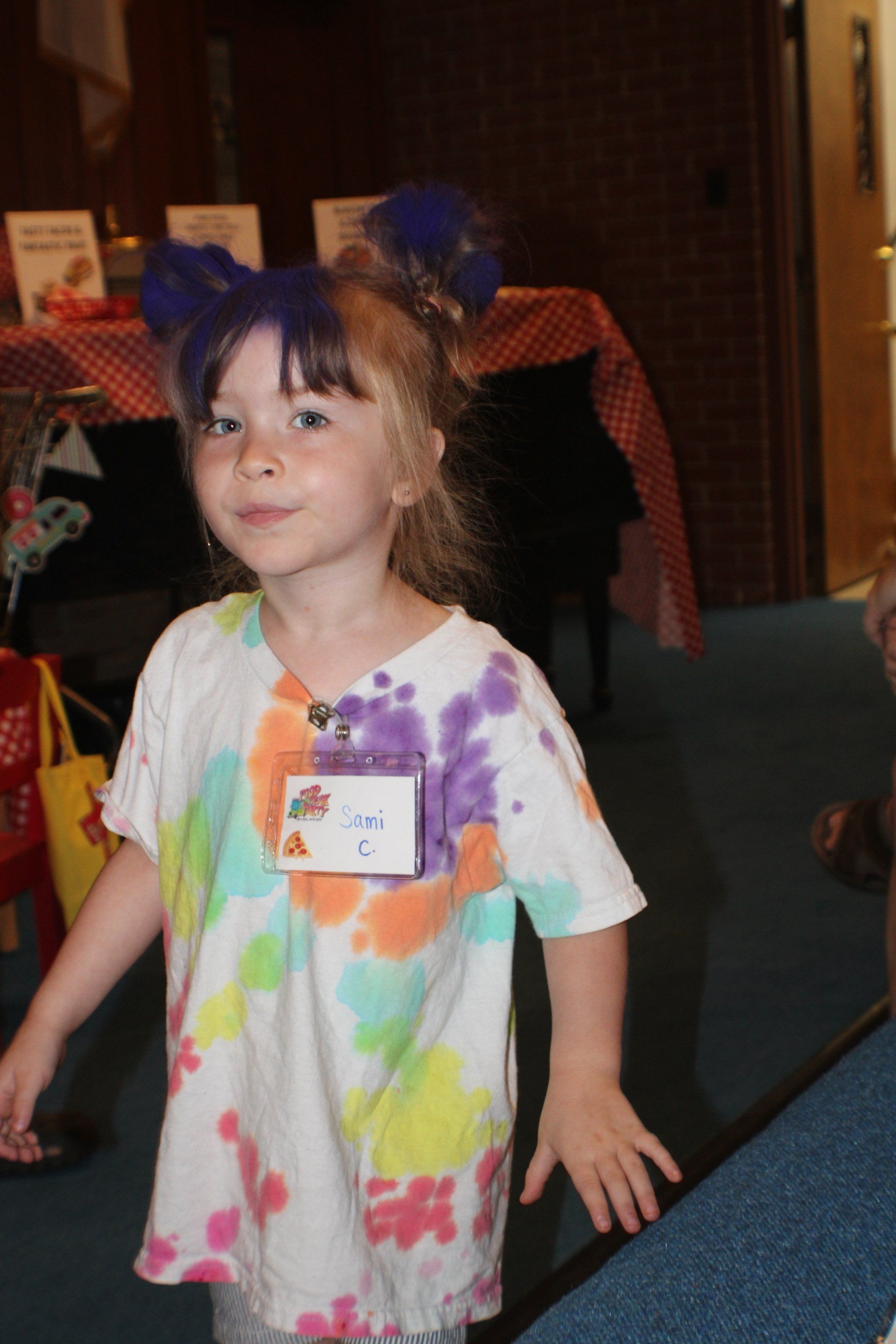 Child in a tie-dye shirt standing indoors, wearing a name tag and smiling at the camera.