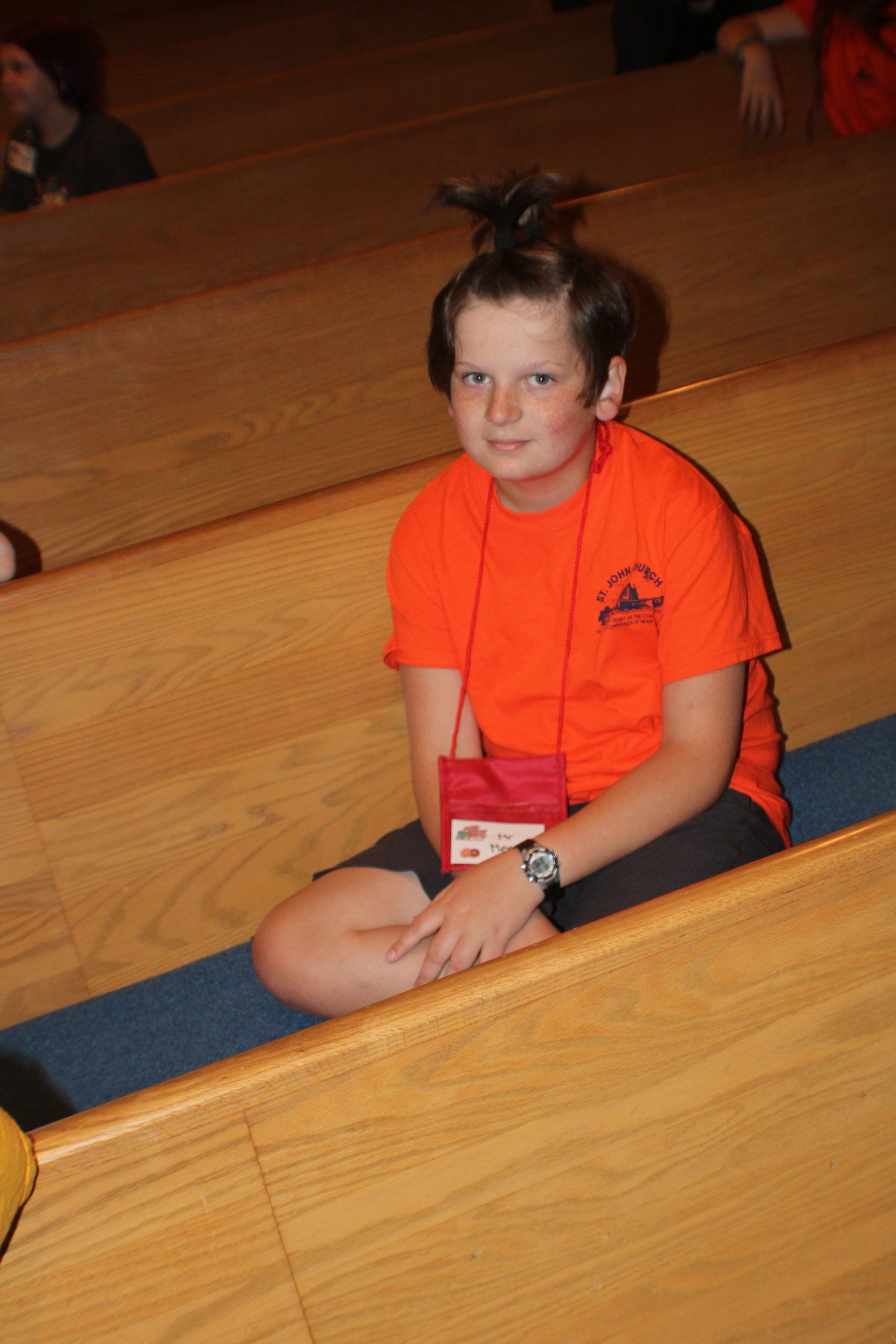 Child in an orange shirt sits cross-legged on a wooden bleacher, looking at the camera.