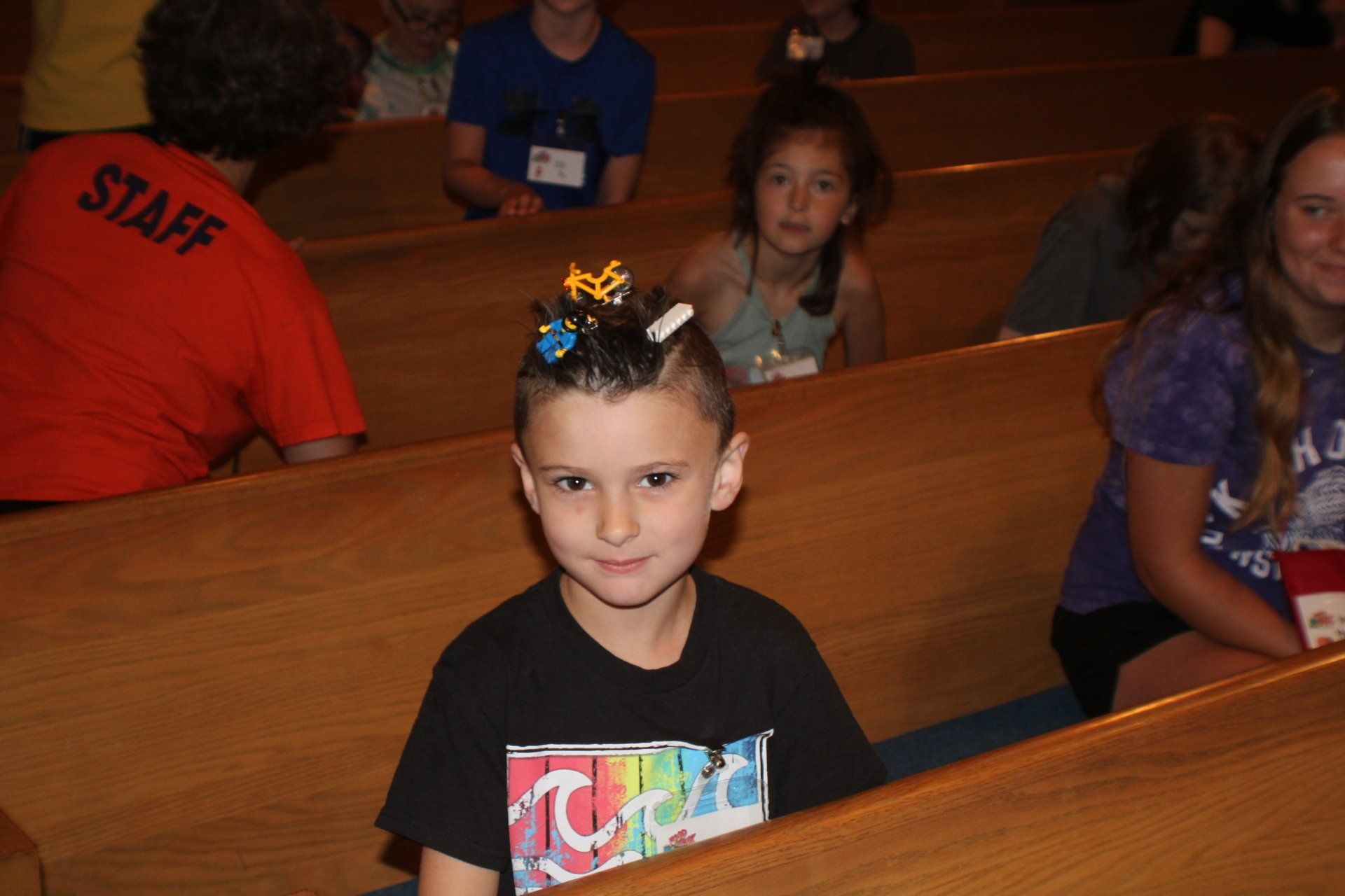 Child with bow in hair sitting in a wooden church pew, holding a colorful card and looking at the camera.