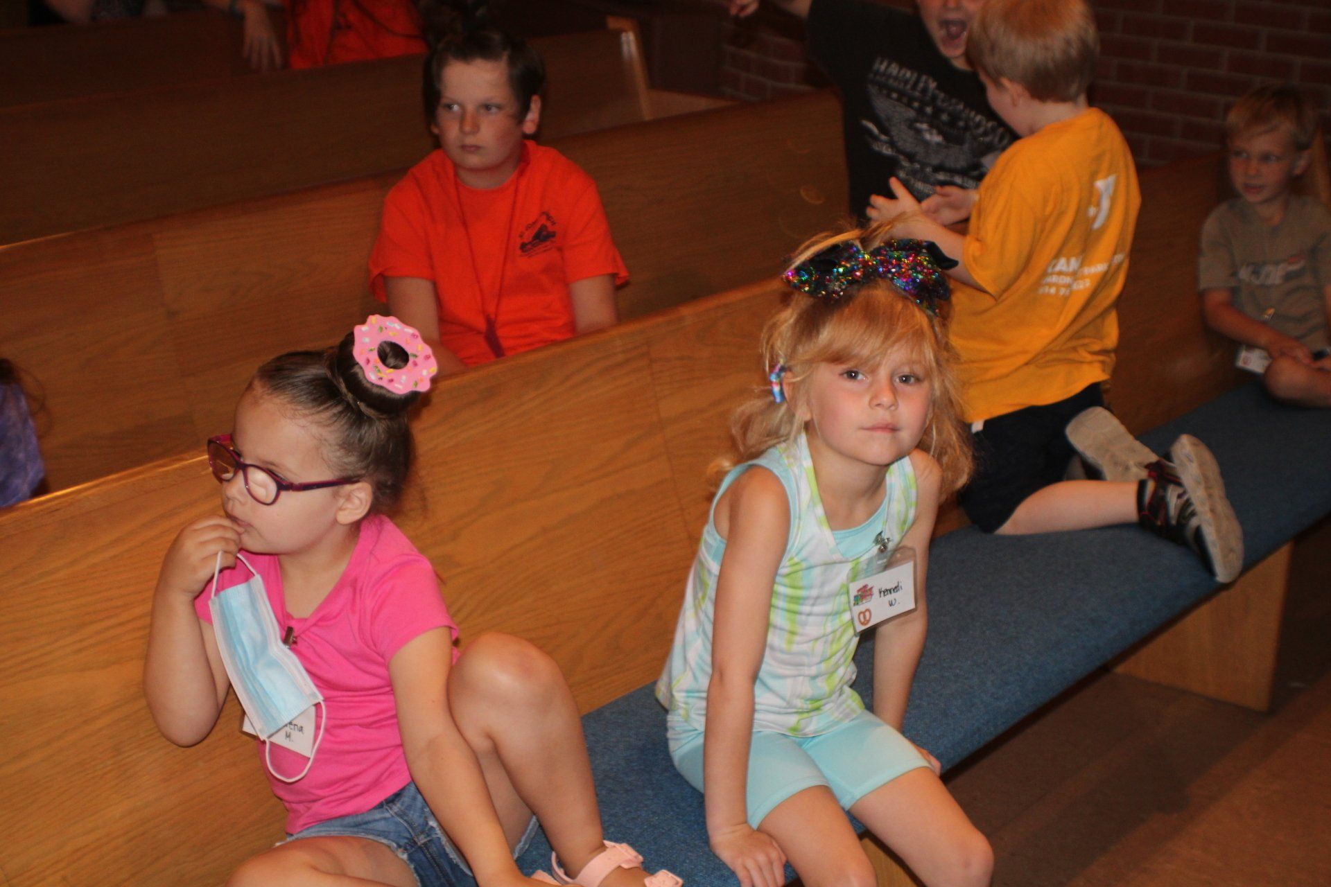 Children sitting on benches in a warm-lit room, with one girl in pink glasses in the foreground