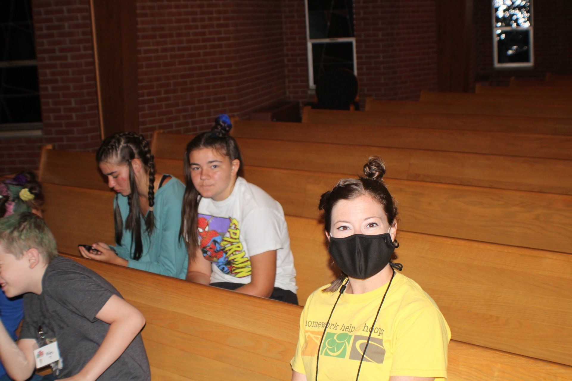 Children sitting on wooden benches in an auditorium, one wearing a black face mask and yellow shirt.