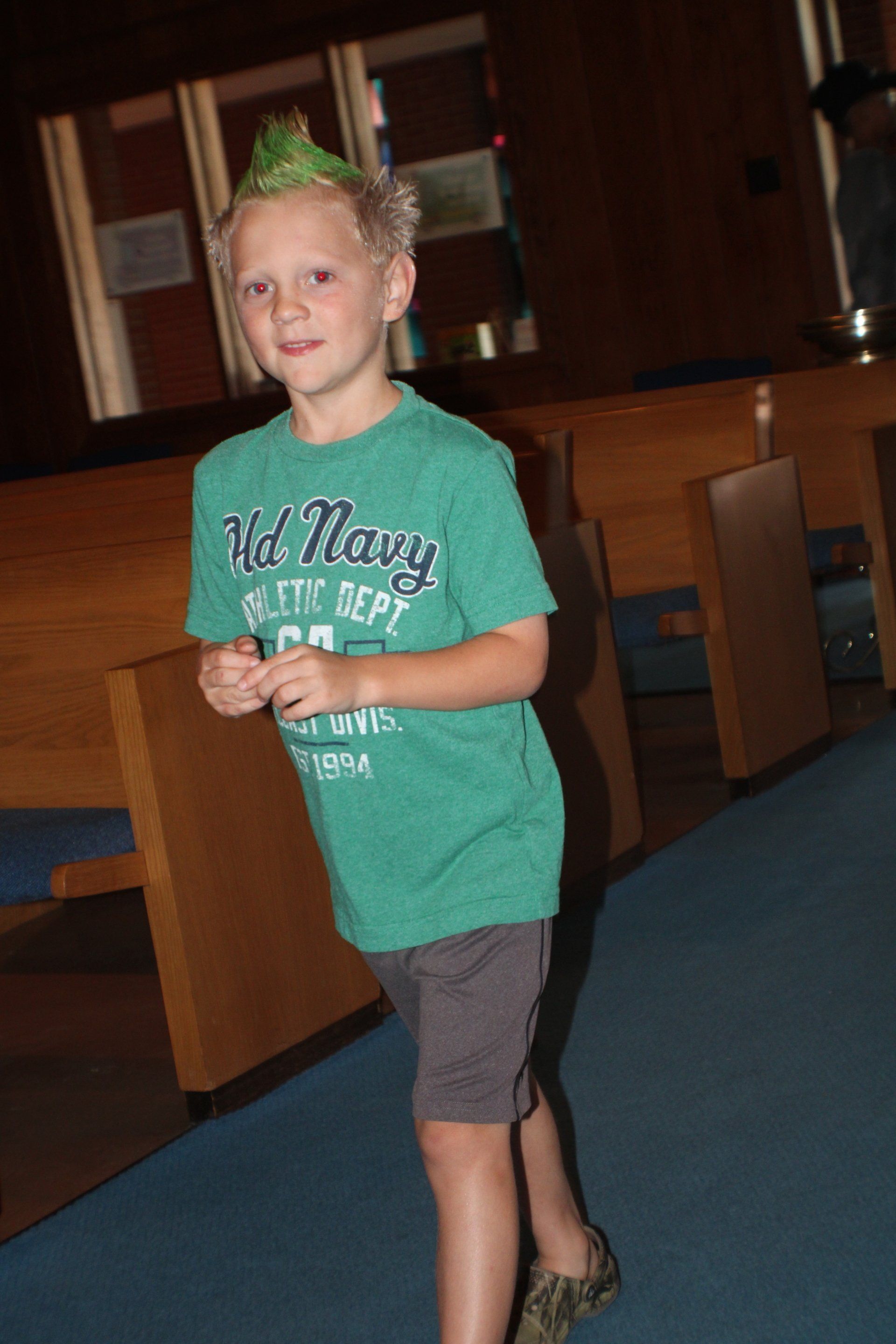 Child with green hair stands indoors in a teal shirt and gray shorts, holding a small object.