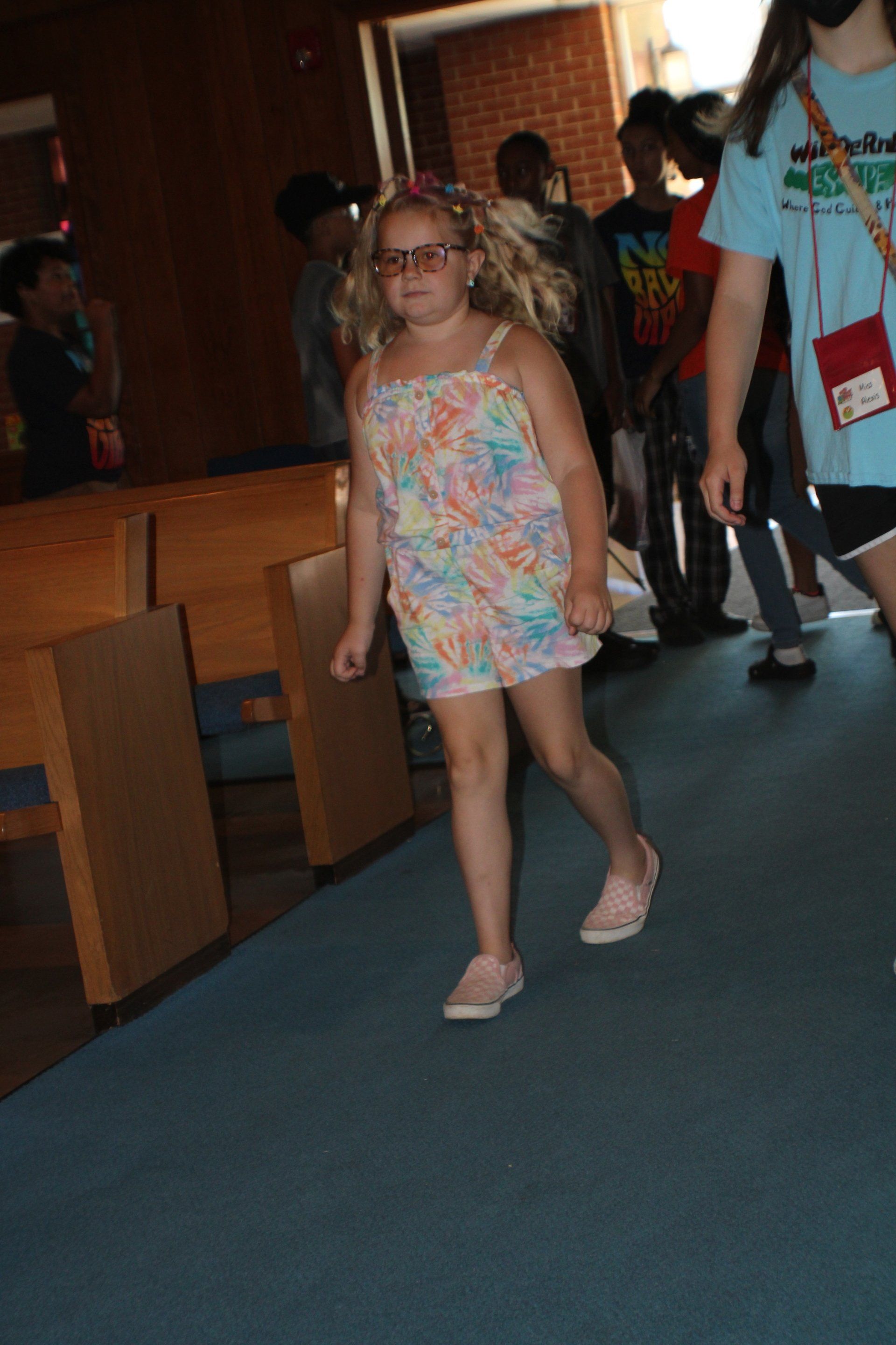 Child walking down a hallway in a colorful dress and glasses, with people in the background.