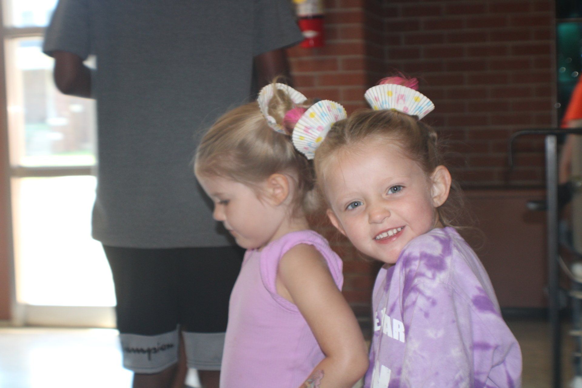 Two children in purple shirts standing indoors, one smiling at the camera.