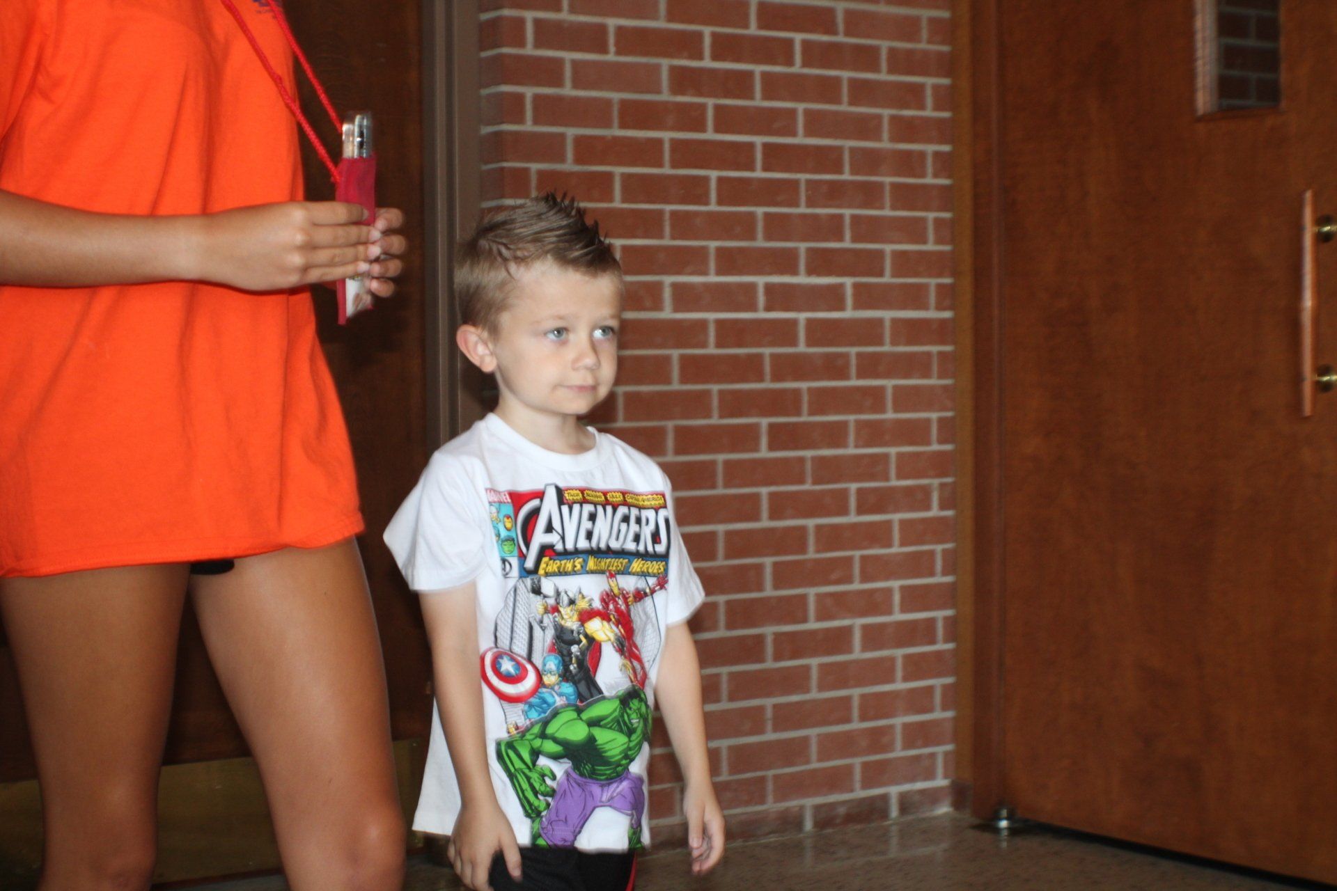 Child standing indoors beside an adult in an orange shirt, near a brick wall and wooden door.