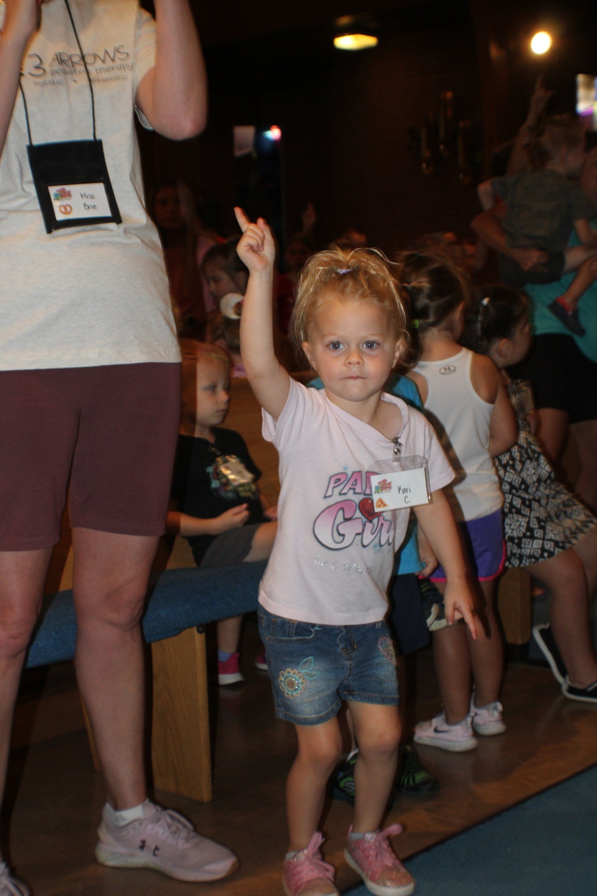 Child in white T-shirt and denim shorts pointing upward at an indoor event, with people in the background