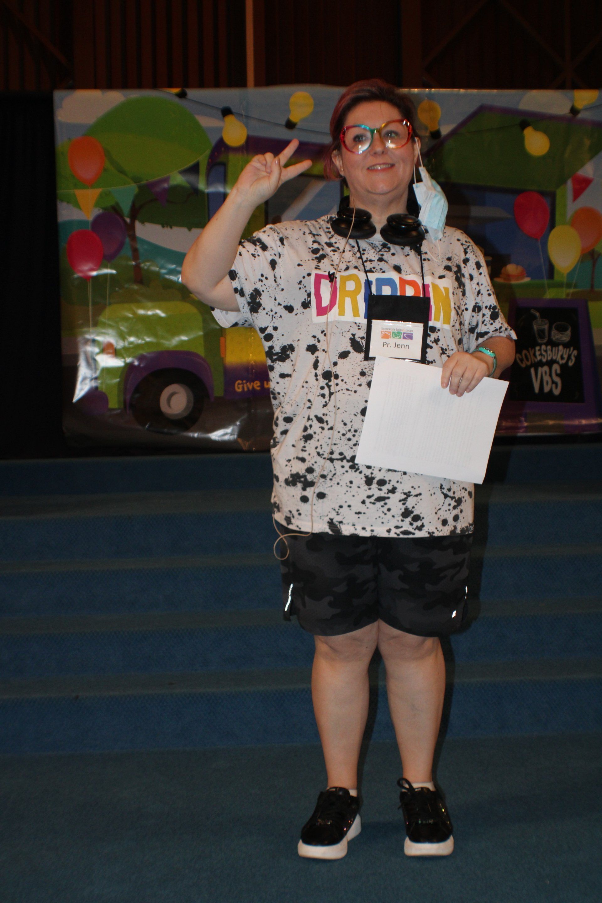 Person standing on a stage, holding a paper and waving, in front of a colorful mural backdrop.