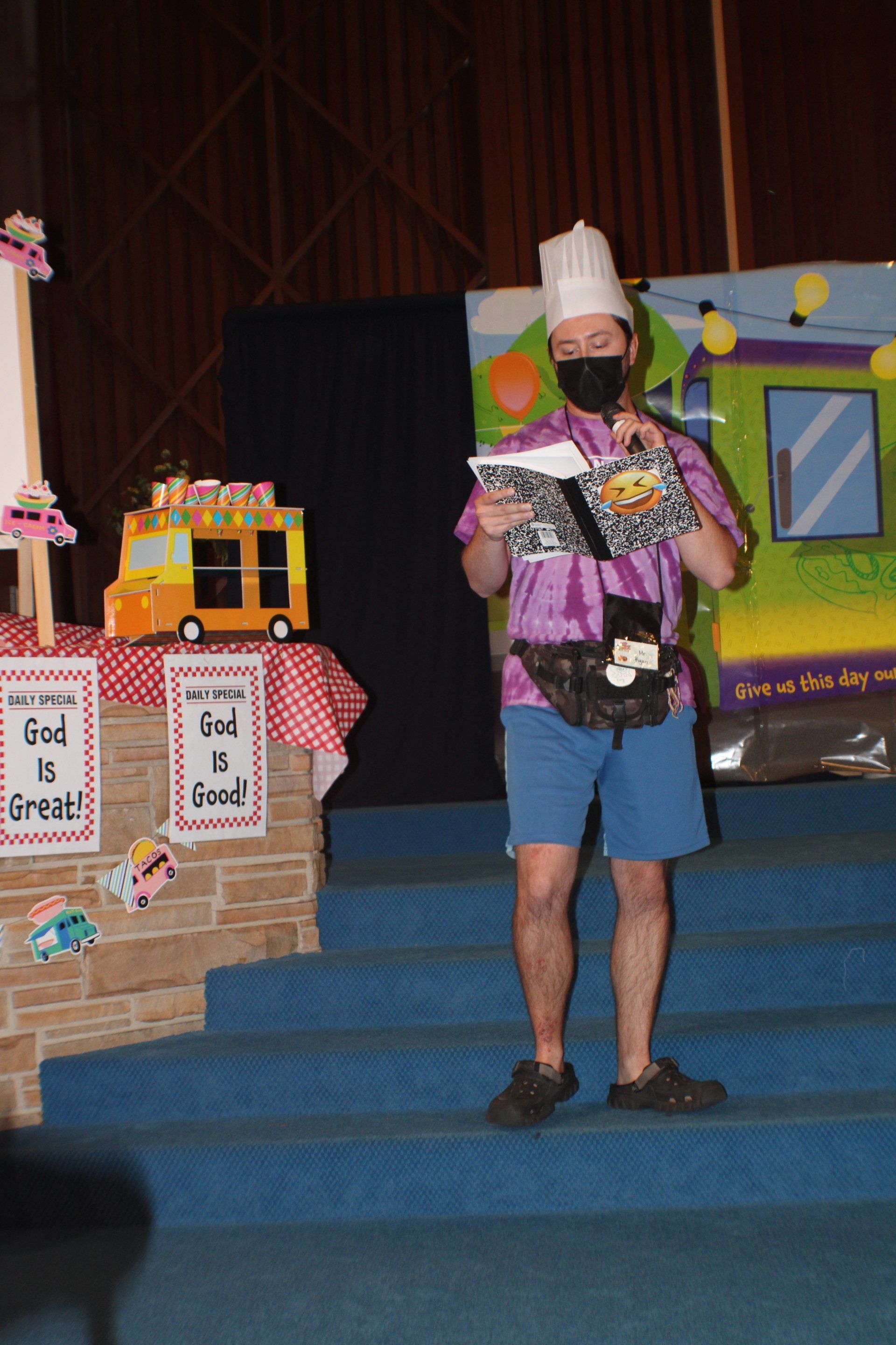 Person in a chef hat stands on stage holding a colorful craft display beside a decorated booth.