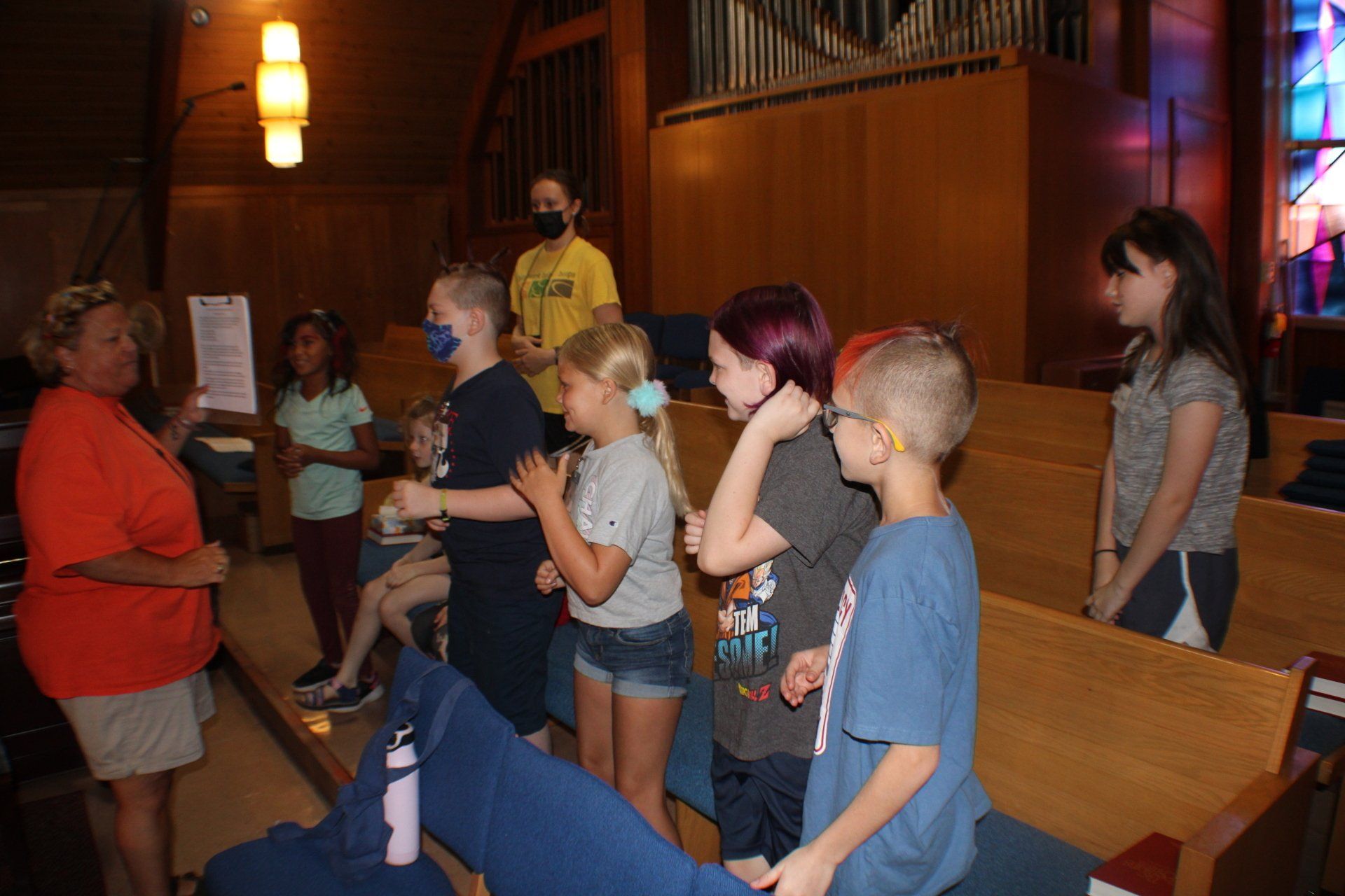Children standing in a church pew area, listening to a woman speaking indoors