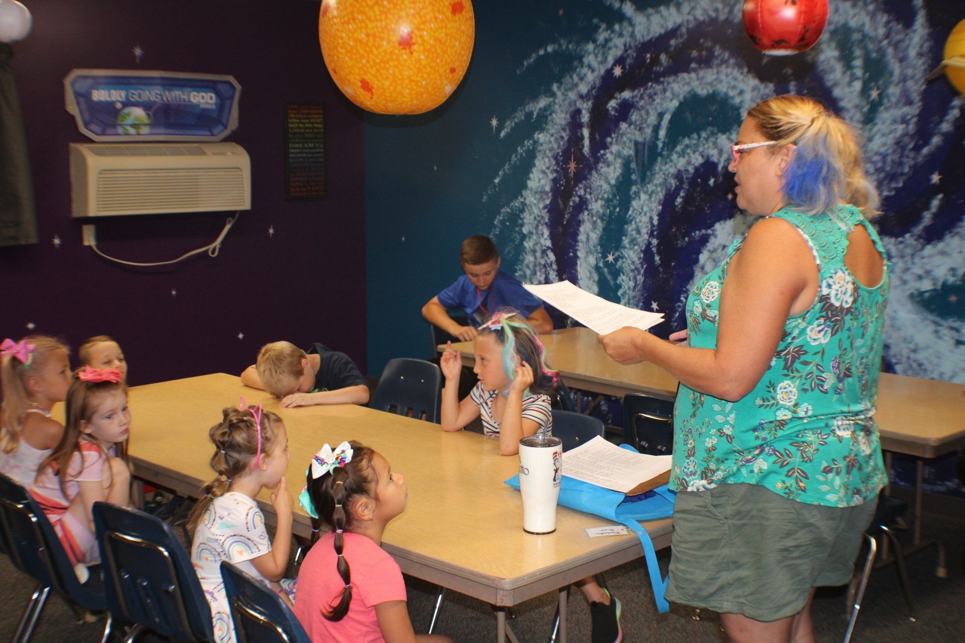 Teacher reading to children in a classroom with a space-themed mural and colorful hanging planets.